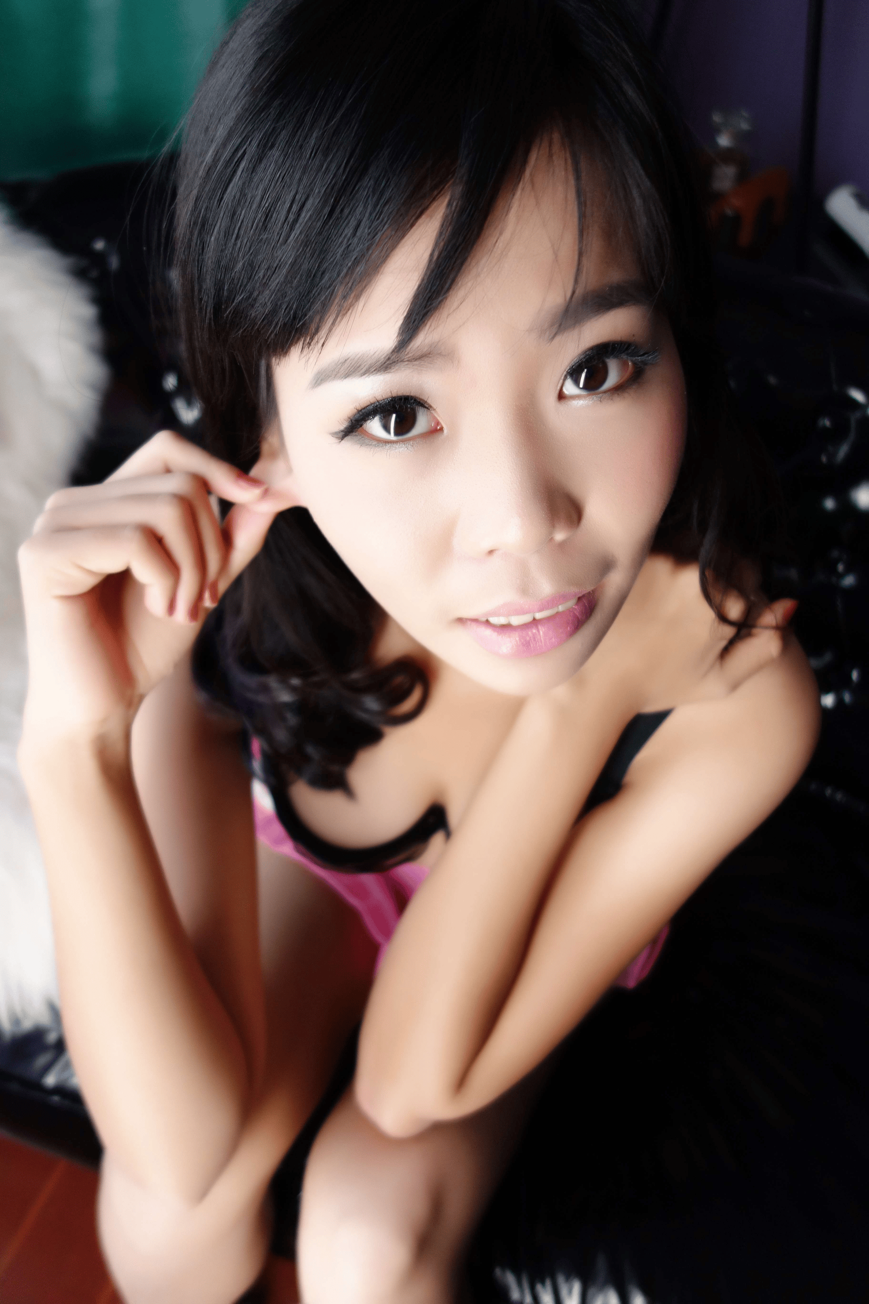 High-angle portrait of a young East Asian woman with wavy dark black hair and side bangs, looking straight up at the camera. She wears light makeup and red nail polish, with one hand gently touching her ear, dressed in a pink camisole with black trim, sitting on a black leather sofa next to a white fuzzy cushion.