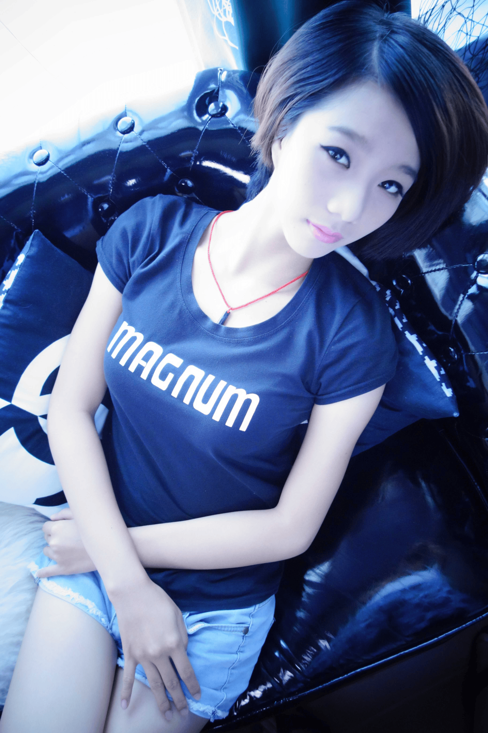 Casual boudoir photo of young short-haired East Asian woman wearing black Magnum graphic t-shirt and cut-off light blue denim shorts seated on tufted black leather sofa.