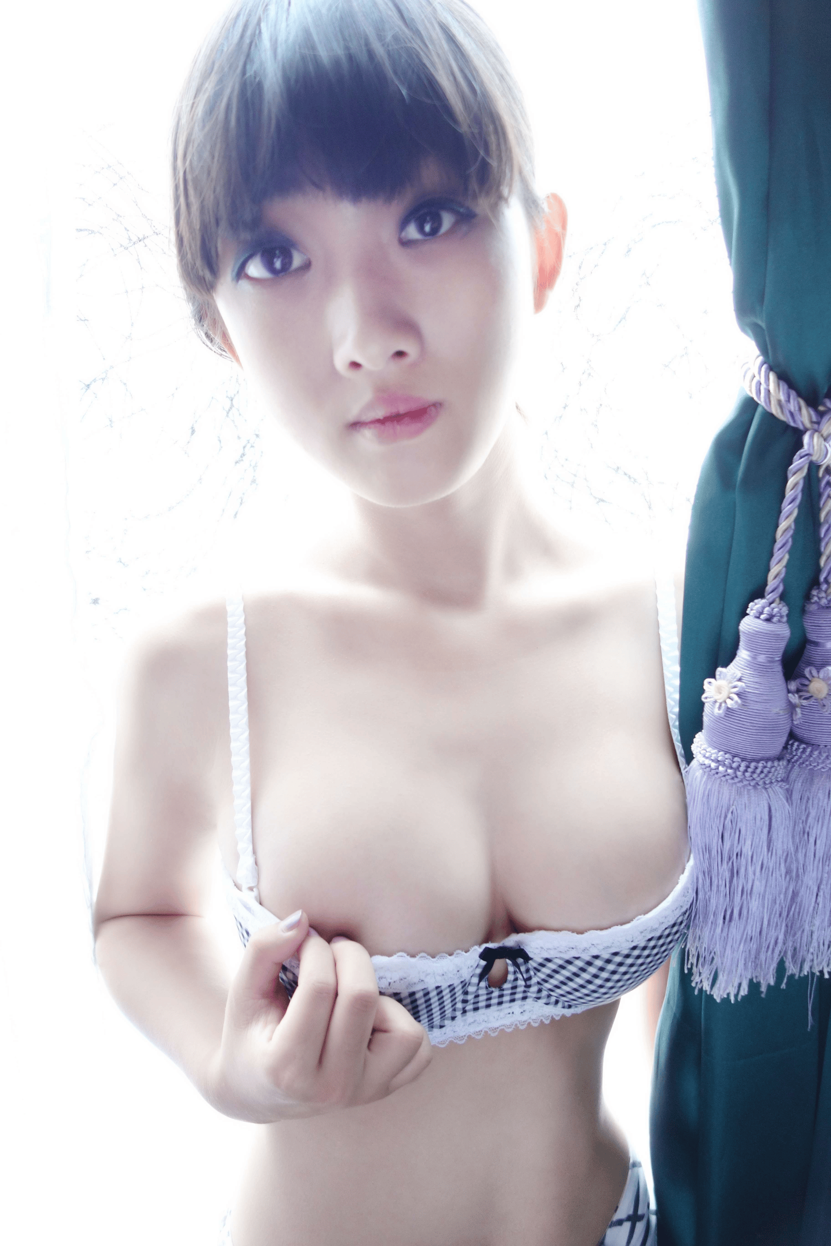 East Asian woman in boudoir photography wearing black and white gingham lace bra, holding bra edge beside dark green curtain with purple tassels near a bright sheer window.