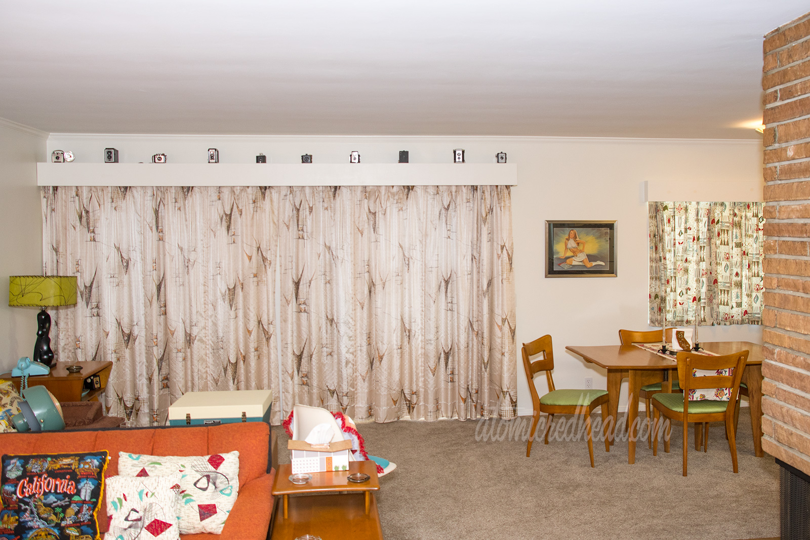 A view into our living room. Along the back wall a long stretch of curtains in tan feature brown asymmetrical shapes. The valance above features multiple vintage cameras.