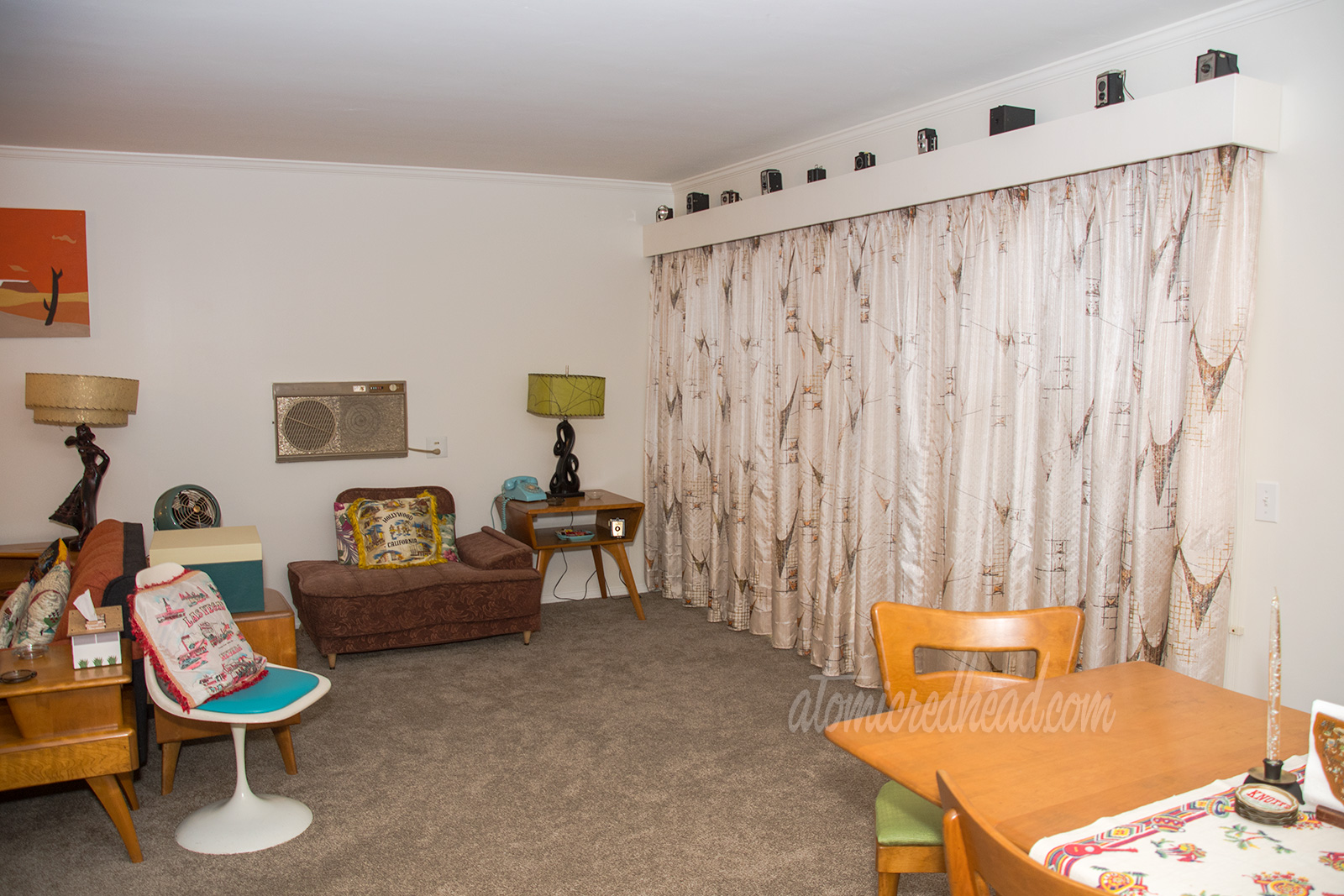 A view into our living room. The curtains hang along the right wall, while an upholstered chair in mauve sits agains the back wall. A maple table features a black modern sculpture style lamp with a lime green shade.
