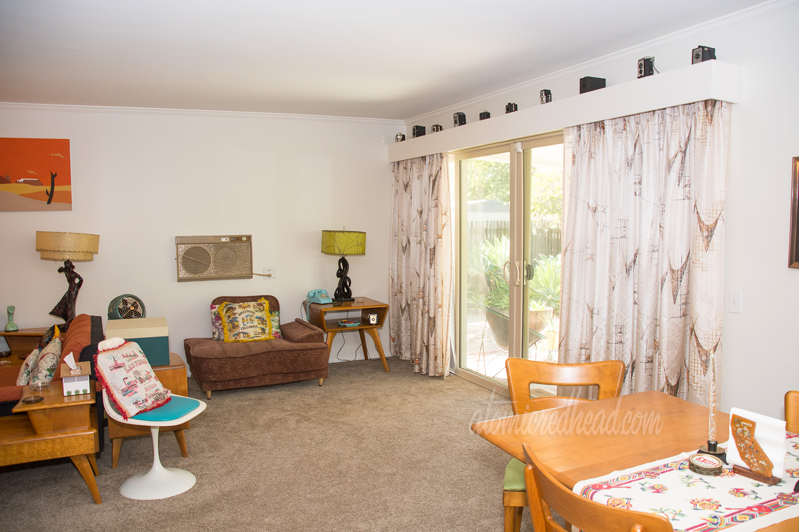A view into our living room. The curtains, open, leading to the patio outside, hang along the right wall, while an upholstered chair in mauve sits agains the back wall. A maple table features a black modern sculpture style lamp with a lime green shade.