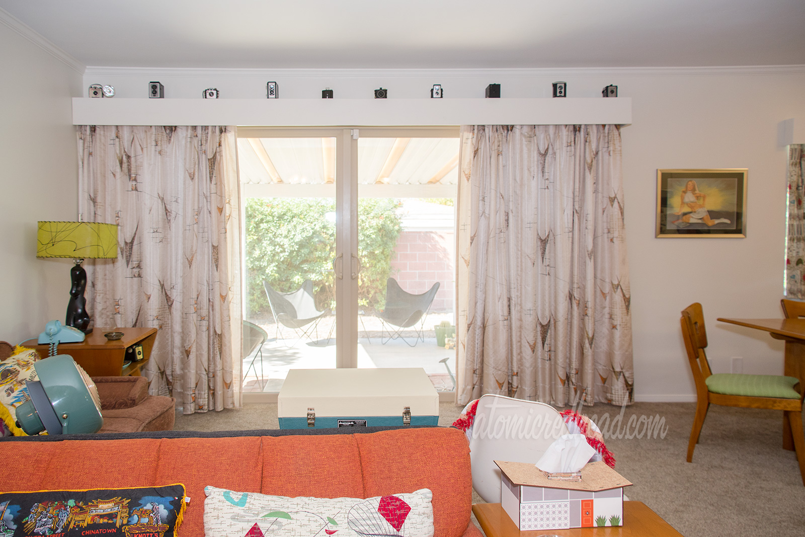 A view into our living room. Along the back wall a long stretch of curtains, open with a view of the backyard, in tan feature brown asymmetrical shapes. The valance above features multiple vintage cameras.