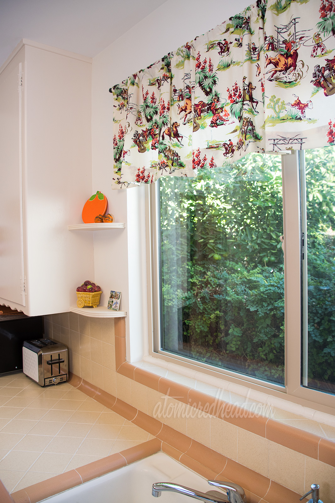 View of the window above the sink in the kitchen, with features a valance of cowboy fabric. Cowboys rest by a fire, some are riding.