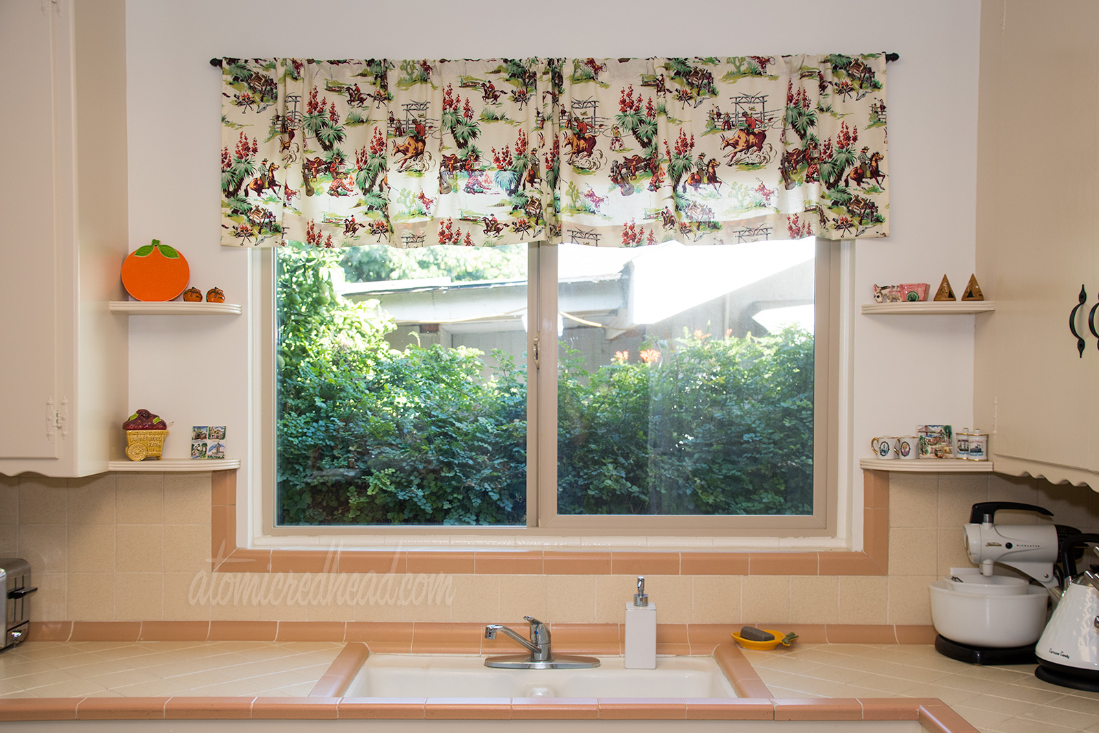 View of the window above the sink in the kitchen, with features a valance of cowboy fabric. Cowboys rest by a fire, some are riding.