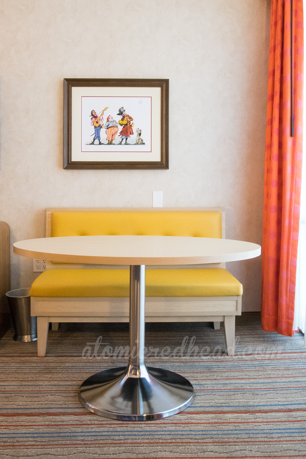 Inside one of the new rooms, stripe carpet, orange curtains, yellow couch, and vintage Pirates of the Caribbean concept art hanging on the wall.