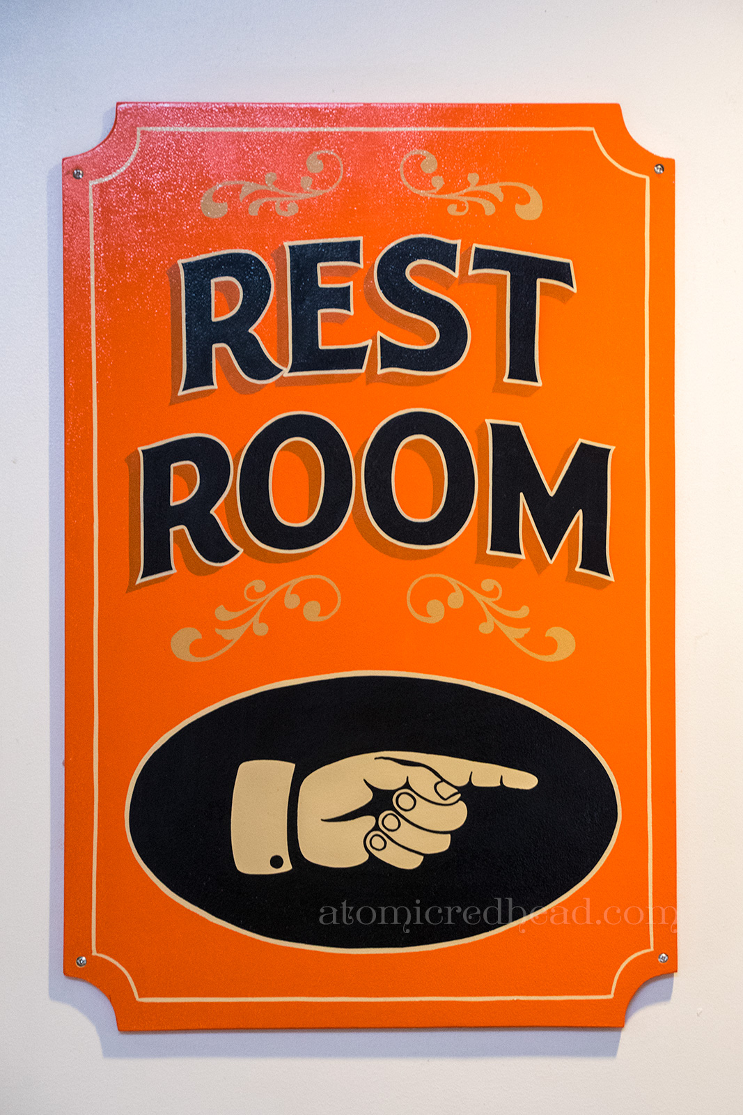 Restroom sign, hand painted, orange, with a finger pointing the way.