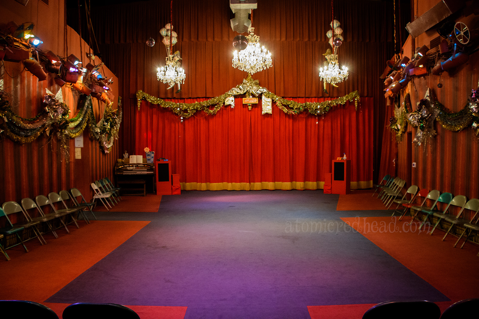The theater inside - red velvet curtains hang all round, three chandeliers hang, gold garlands trim the edge.