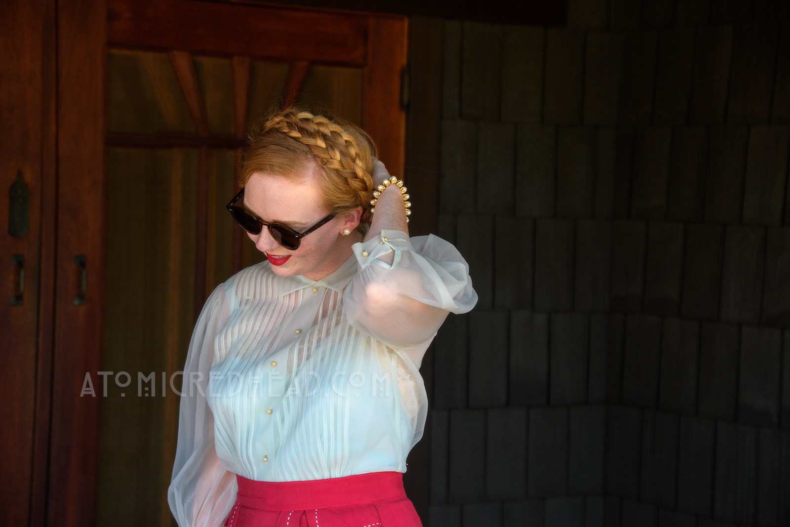Standing on the back porch of the Gamble House, wearing a pale blue sheer blouse and magenta skirt with white trim, and a brown woven basket purse.