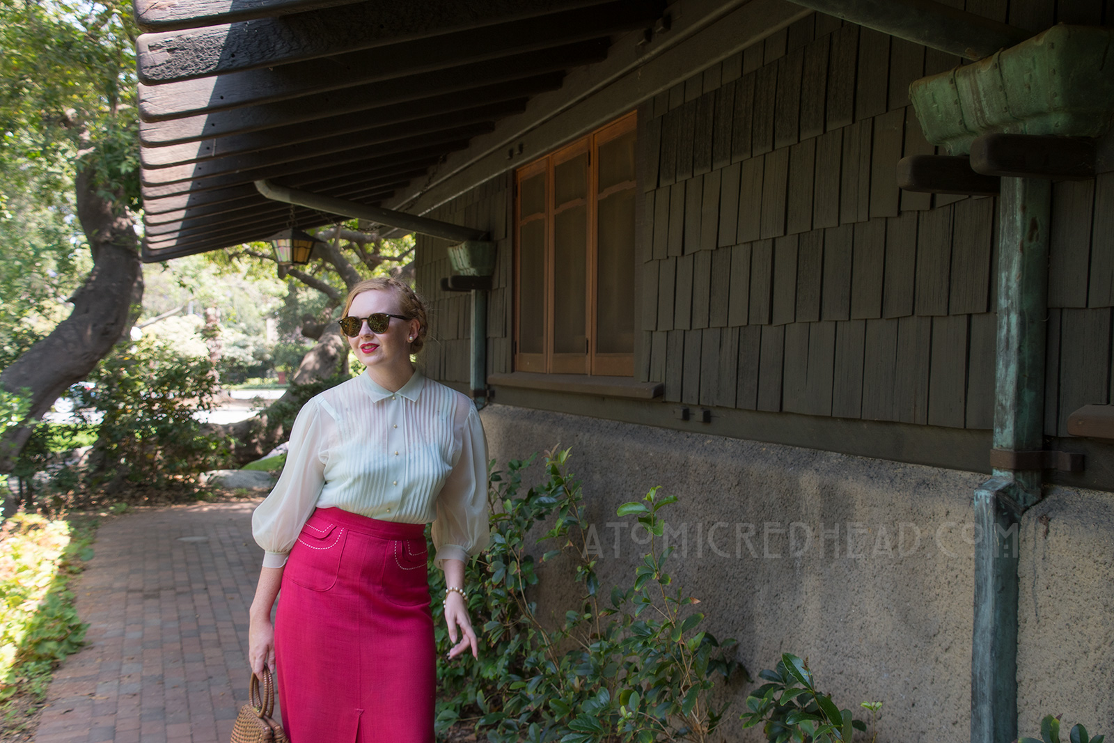 Standing near the garage of the Gamble House, wearing a pale blue sheer blouse and magenta skirt with white trim, and a brown woven basket purse.