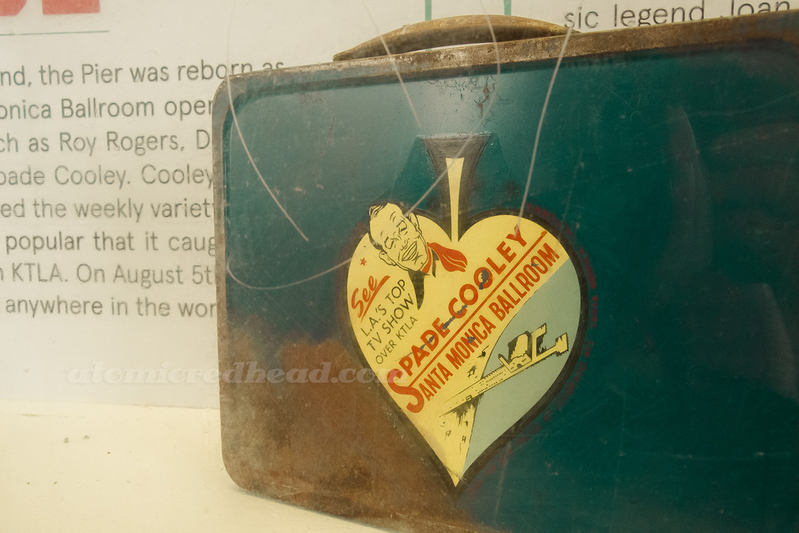 A vintage lunchbox featuring a cartoon of Spade Cooley, along with an illustration of the pier. 