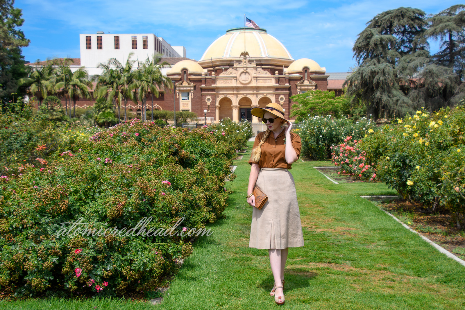 Me, outside of the museum in the rose garden, wearing a straw hat in brown velvet trim, a brown blouse with a gold and green brooch featuring the face of King Tut, a tan linen skirt, and brown and tan shoes.