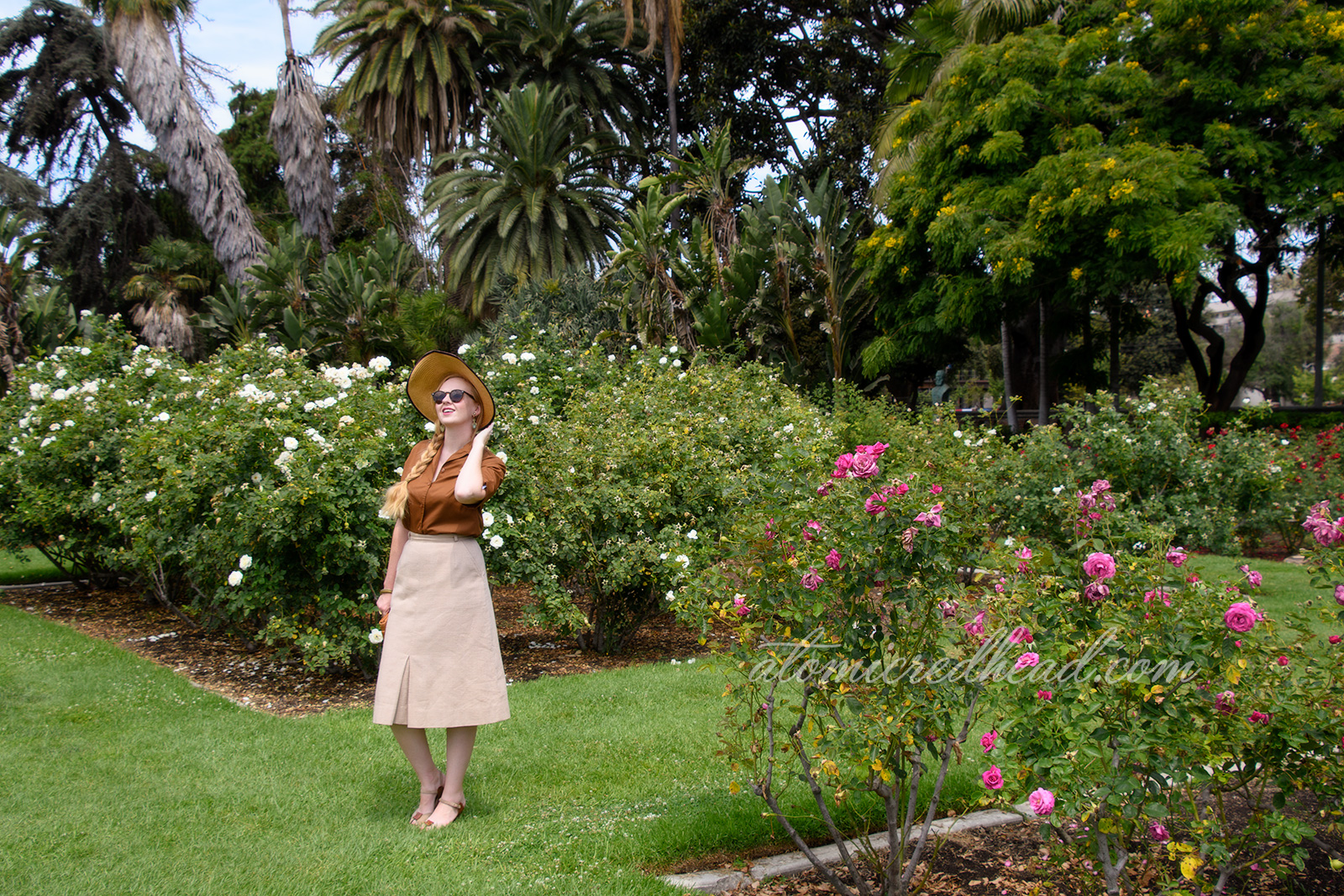 Me, outside of the museum in the rose garden, wearing a straw hat in brown velvet trim, a brown blouse with a gold and green brooch featuring the face of King Tut, a tan linen skirt, and brown and tan shoes.