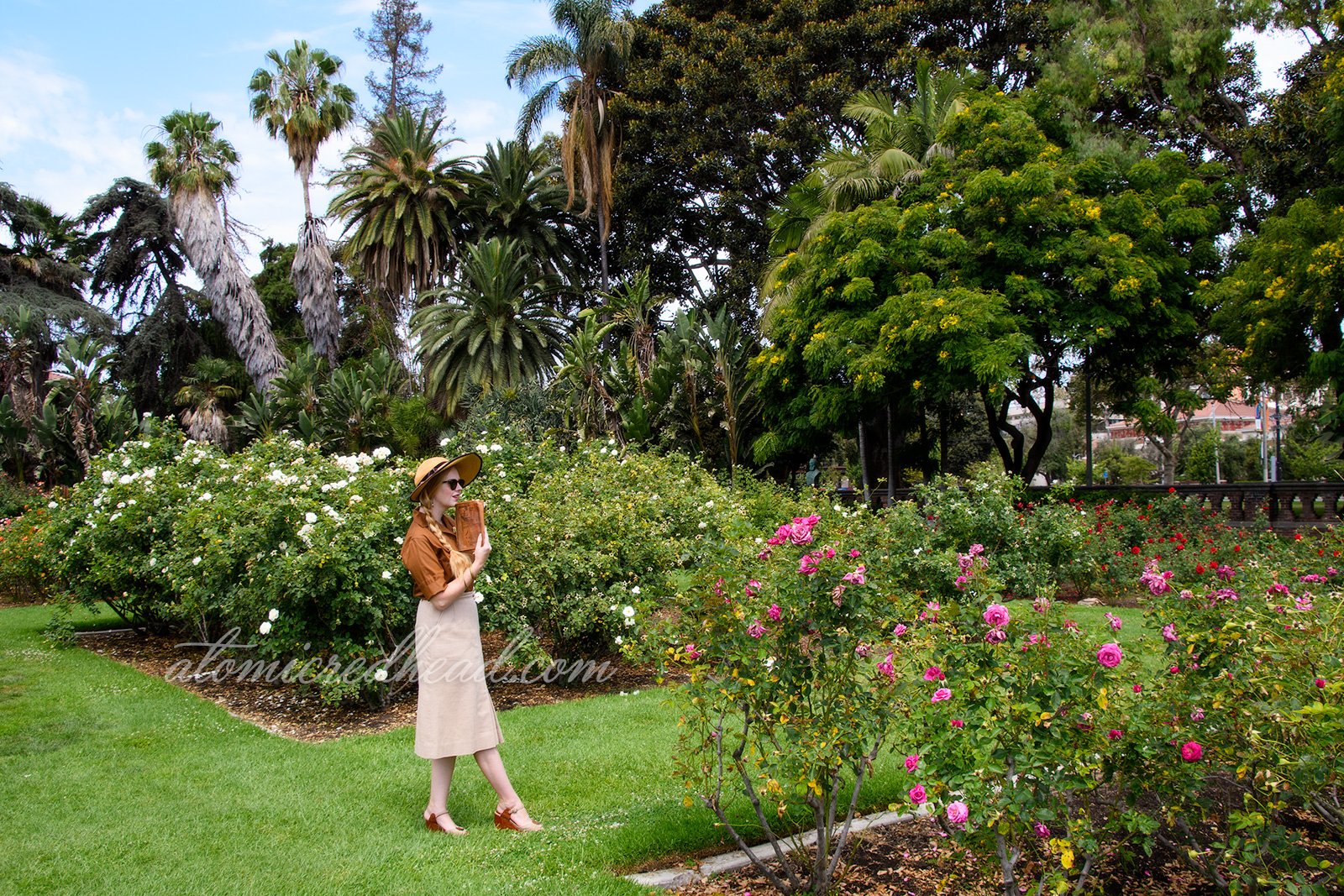 Me, outside of the museum in the rose garden, wearing a straw hat in brown velvet trim, a brown blouse with a gold and green brooch featuring the face of King Tut, a tan linen skirt, and brown and tan shoes.