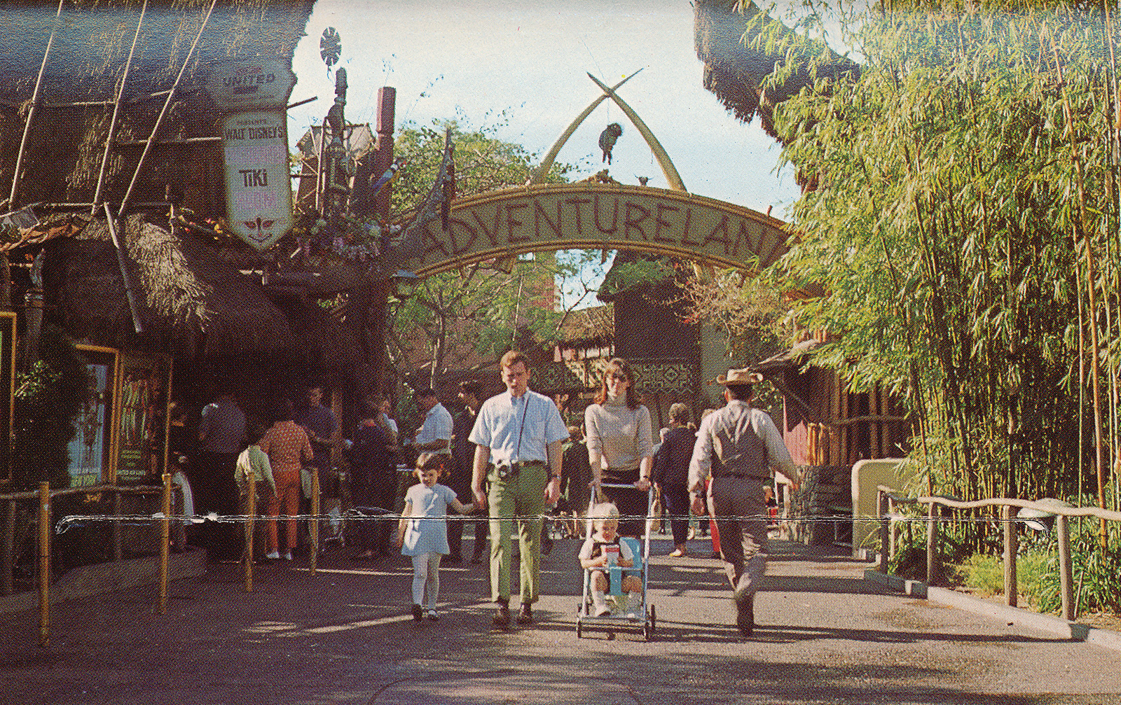 The entrance to Adventureland, a combination of tiki and African culture merge together. Crossed elephant tusks stand above the text reading "Adventureland" that is spelled out in wood. The Enchanted Tiki Room off to the left.