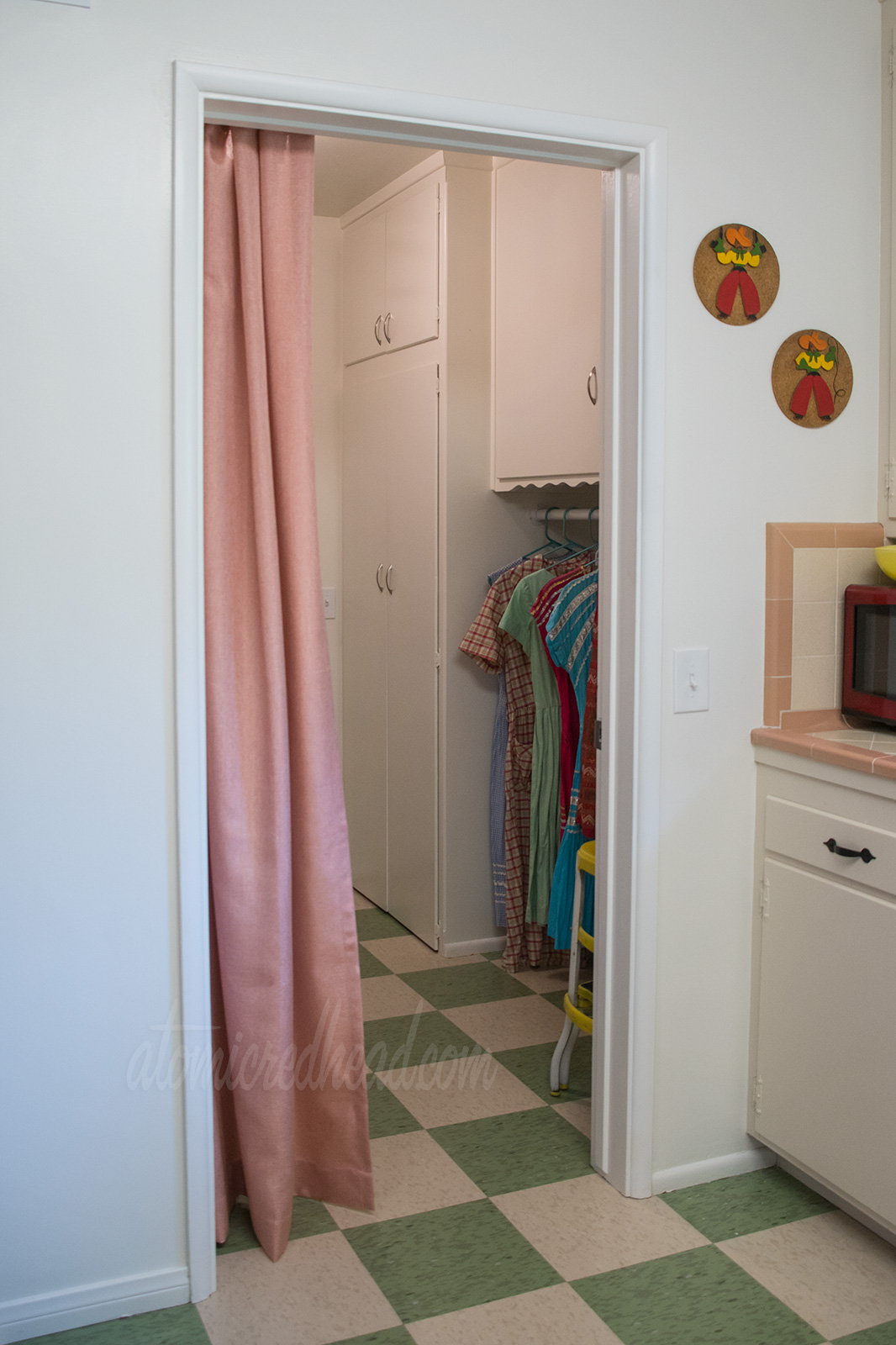 A look into the laundry room from the kitchen. Green and pale yellow check flooring extend into the laundry room. Pale coral curtains hang in the doorway to the left, and just peeking from the laundry room a clothing bar hold clothing that is hanging to dry.