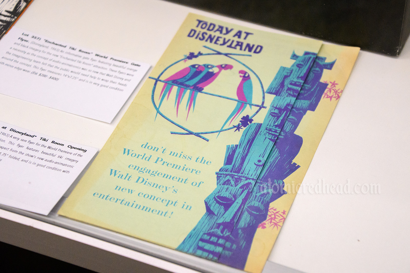 Paper piece advertising the grand opening of the Enchanted Tiki Room, white, with a purple and green tiki totem on the right, and birds on the left.