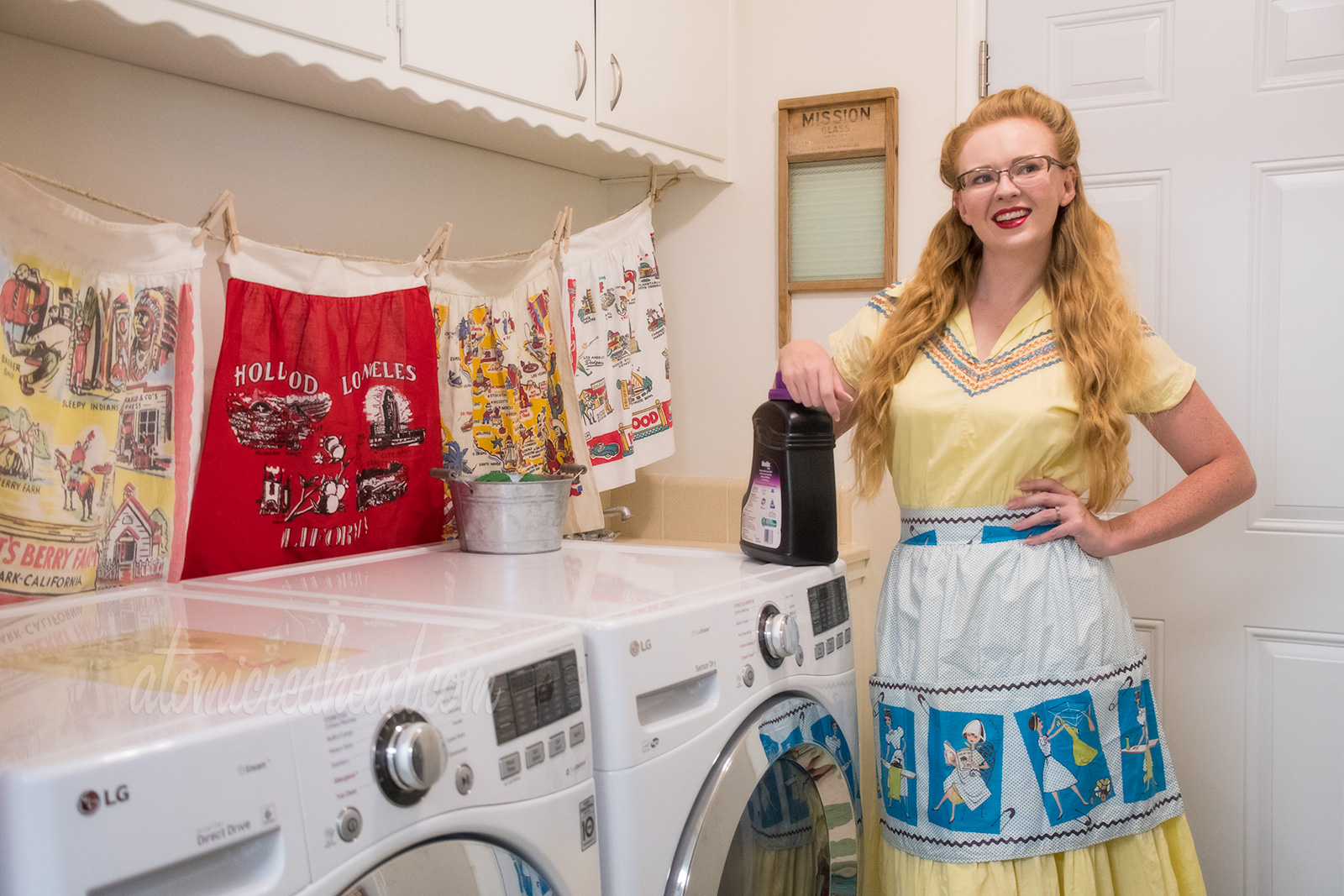 Standing in the laundry room, wearing a yellow dress with blue trim and a blue and white apron featuring a cartoon illustration of a woman doing laundry and getting her hair done.