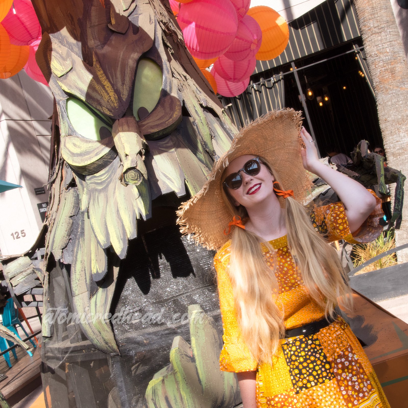 Myself standing in front of a spooky tree float, wearing a patchwork print dress of orange, yellow, black and white, as a large straw hat.