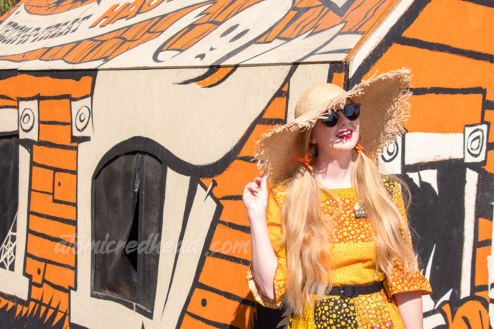 Myself standing in front of a float made to look like a vintage sucker box that also looks like a haunted house, wearing a patchwork print dress of orange, yellow, black and white, as a large straw hat