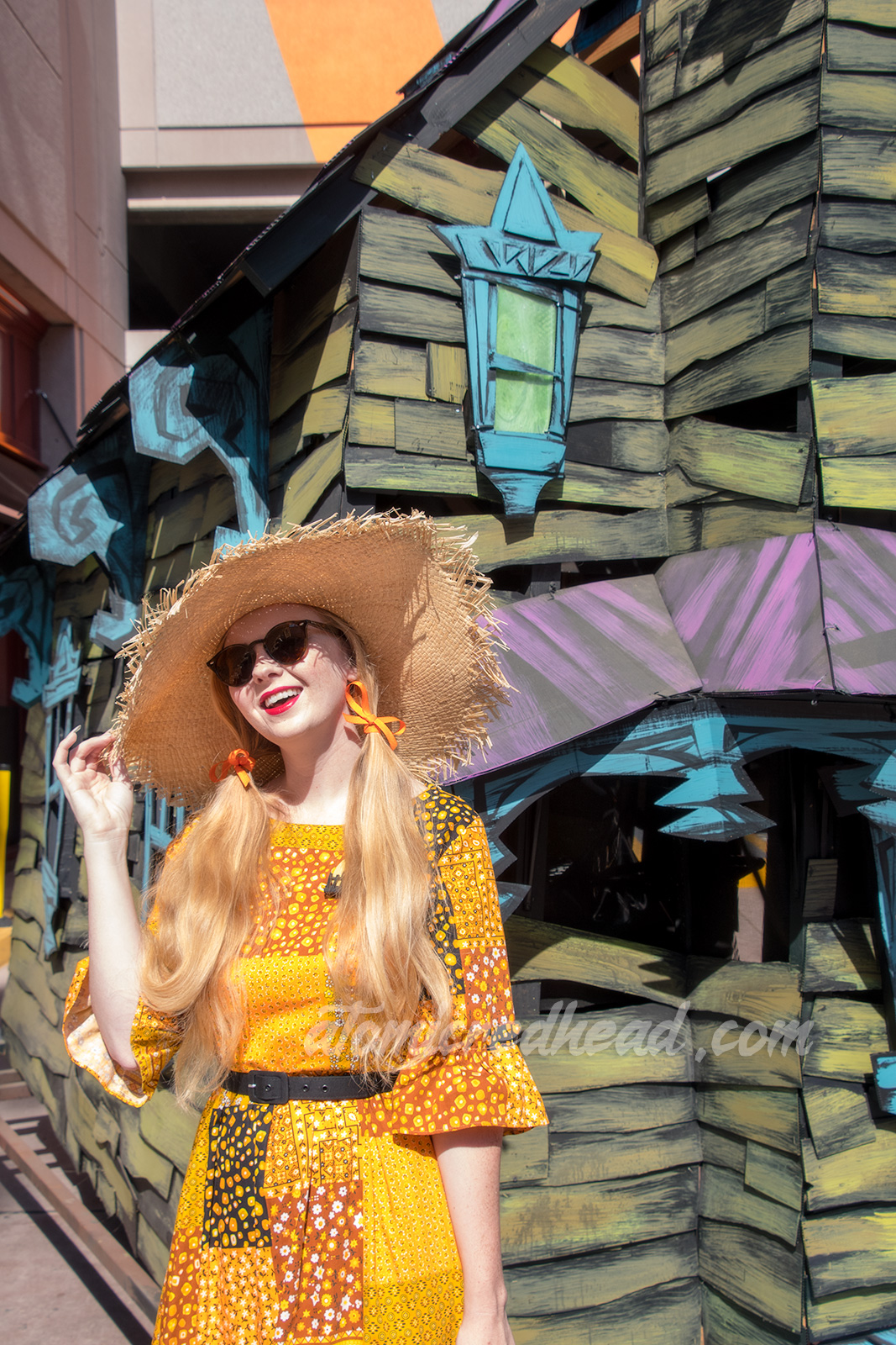 Myself standing in front of a green, purple, and blue haunted house float, wearing a patchwork print dress of orange, yellow, black and white, as a large straw hat