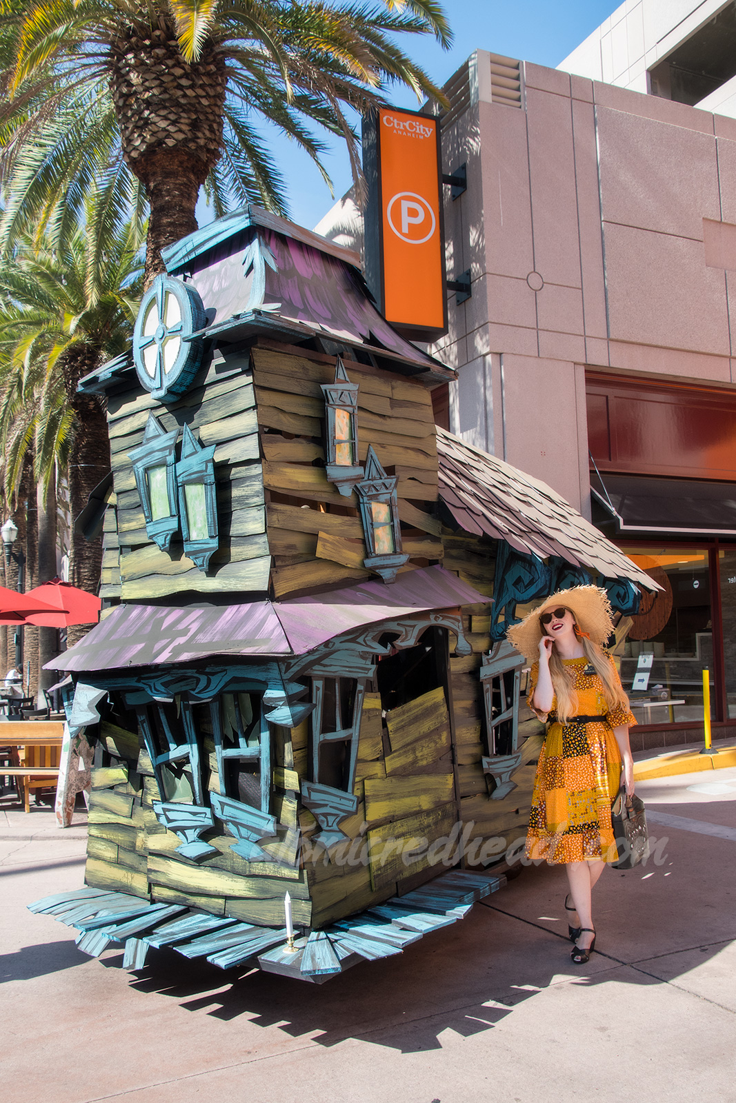 Myself standing in front of a green, purple, and blue haunted house float, wearing a patchwork print dress of orange, yellow, black and white, as a large straw hat