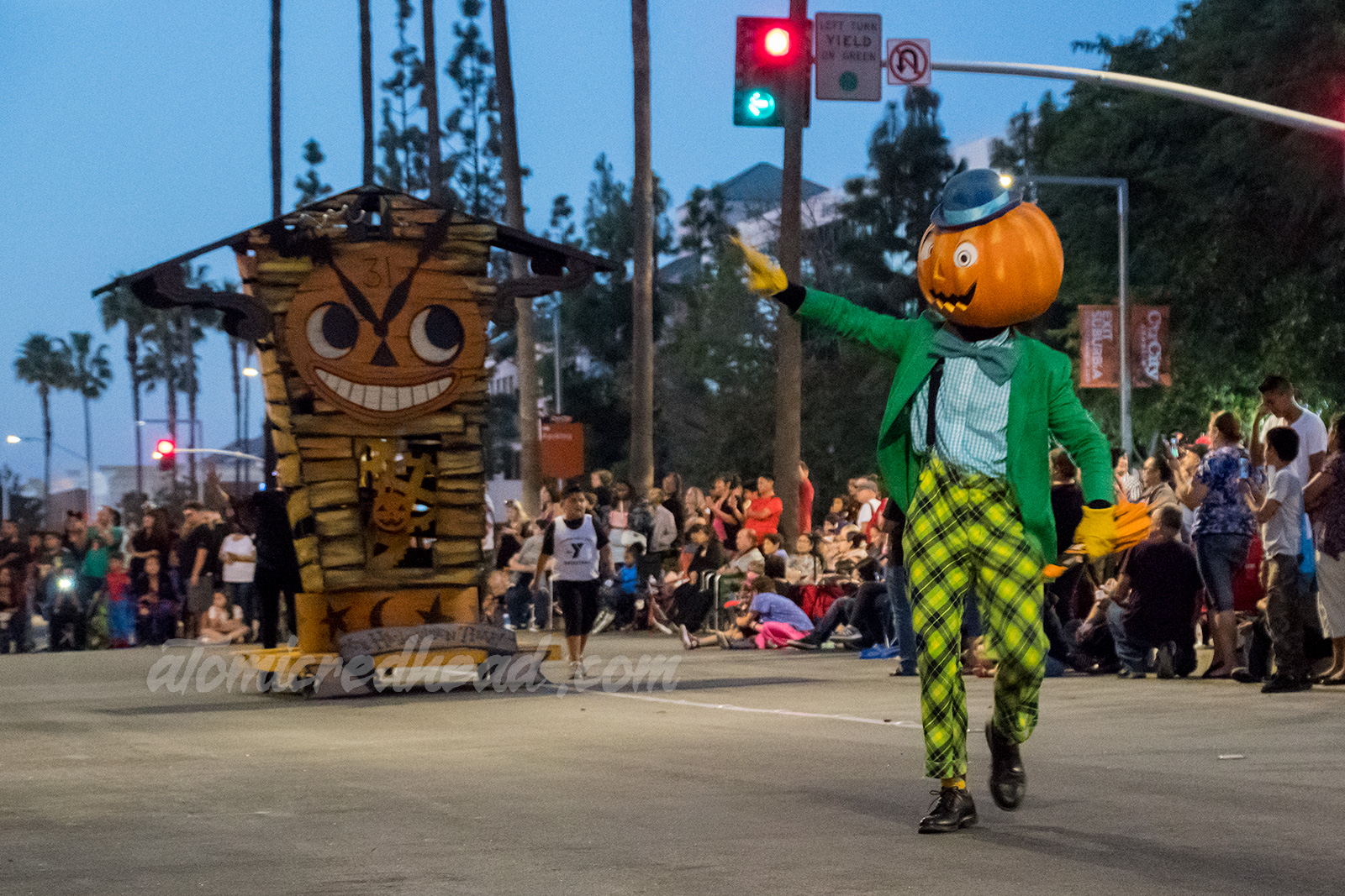 A parade performer wears a green and yellow costume and a large jack o'lantern head with a blue bowler on top.
