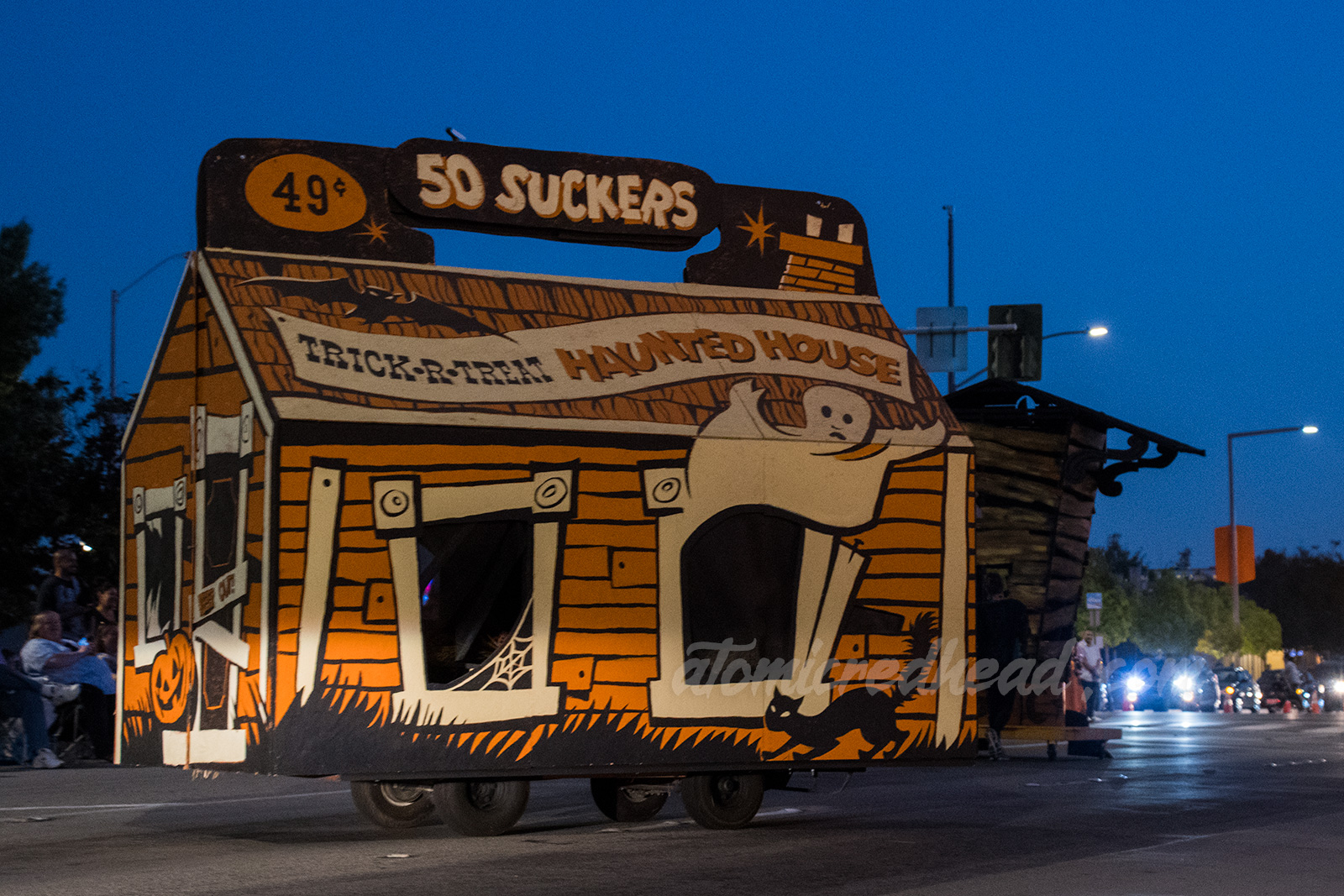 A orange, black, and white, float that is made to look like an old sucker box, and is also a haunted house, with a ghost floating out a window.
