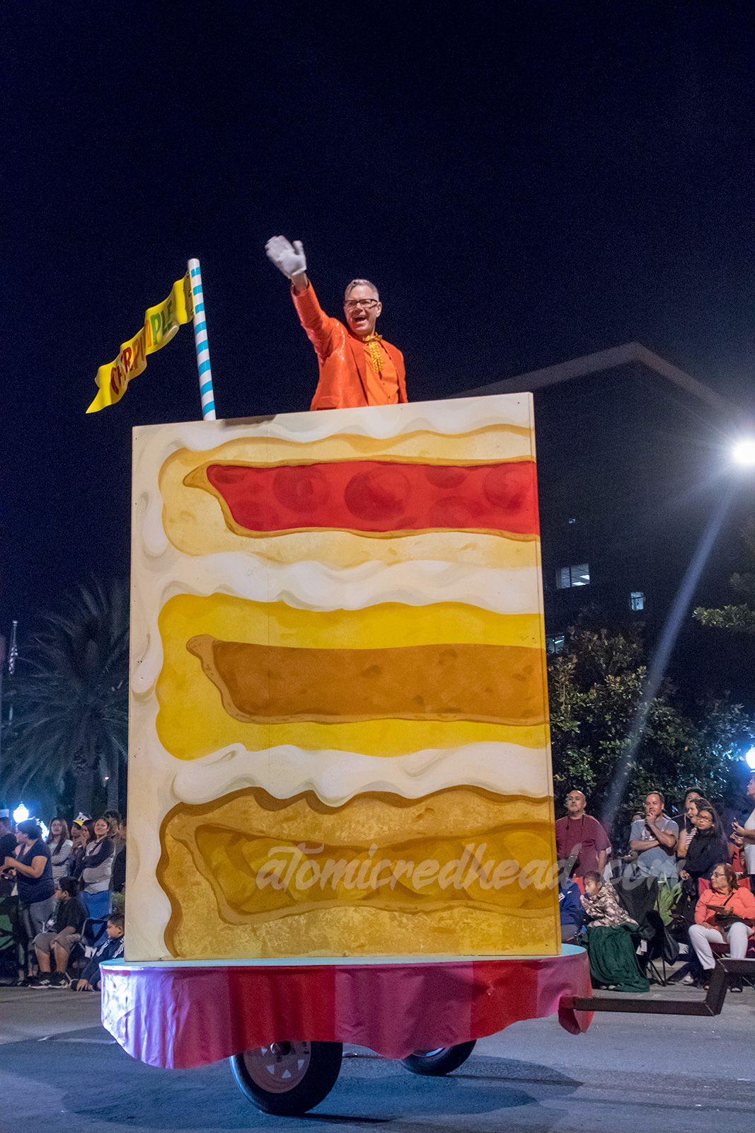 Charles Phoenix sits atop a slice of his "Cherpumple" cake, which is a cherry, pumpkin, and apple pie stacked on top of each other, baked into a yellow cake.