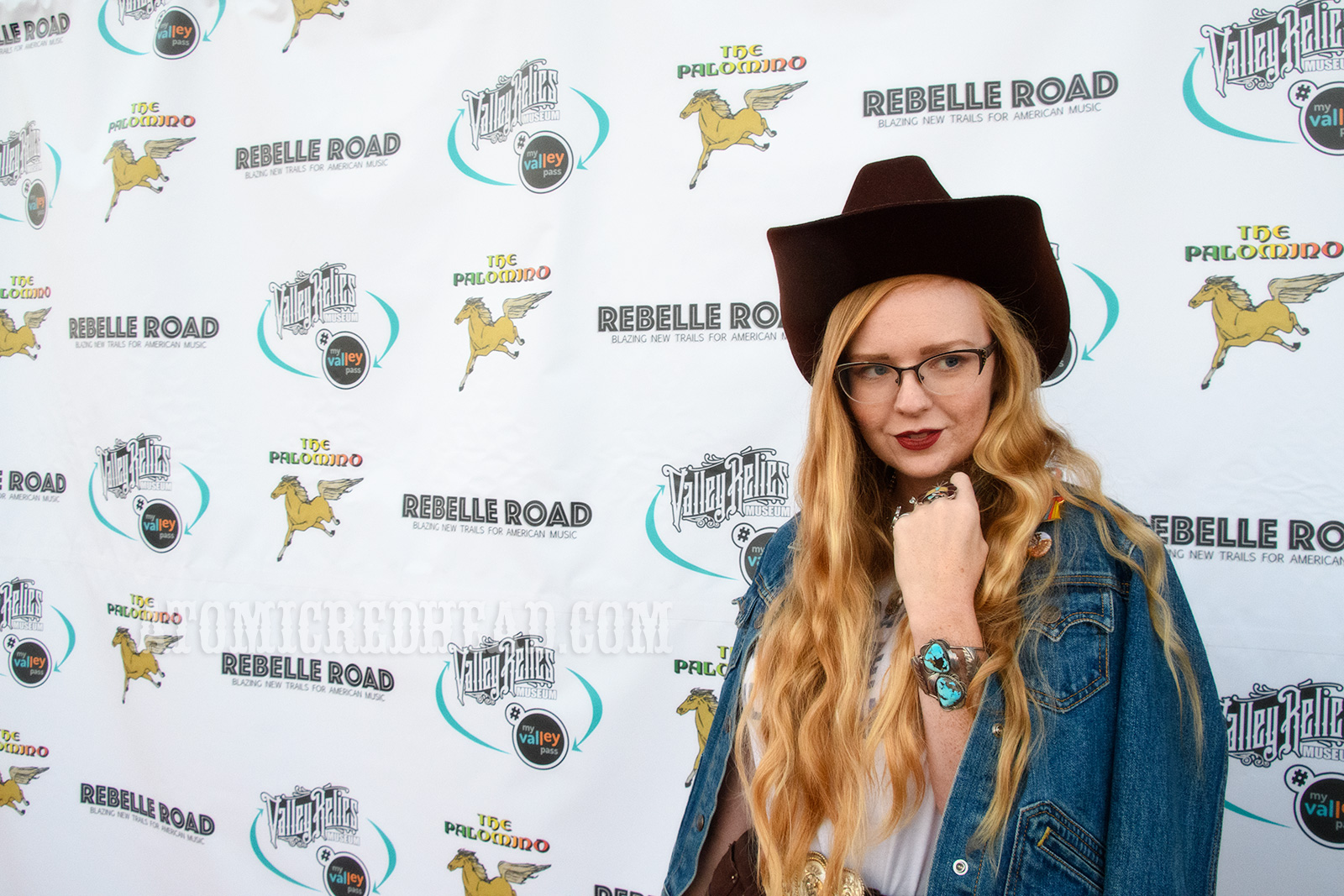 Myself, standing in front of a backdrop featuring the Palomino logo of a horse, and text for the event company, Rebelle Road, and the museum Valley Relics, wearing a blue jean jacket, white t-shirt reading "Gram Parsons and the Fallen Angels" and a dark brown cowboy hat.