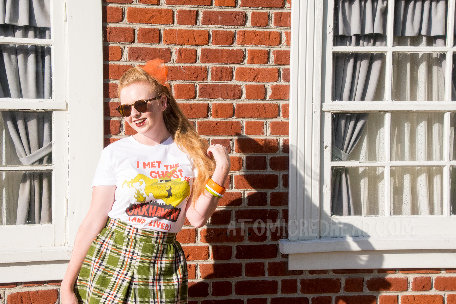 Myself, standing in front of a brick wall, wearing a white t-shirt with an image of a green ghost in a witch hat, rising from a silhouette of a town. Orange text reads "I met the Ghost of Oakhaven (and lived)," a skirt in matching green, orange, white and black, and an orange scarf in my pony tail.