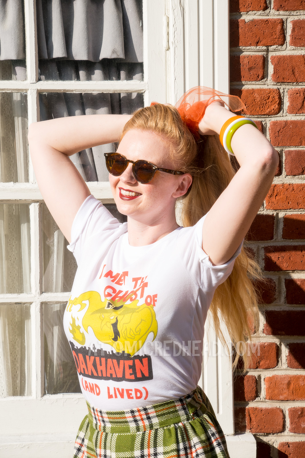 Myself, standing in front of a brick wall, wearing a white t-shirt with an image of a green ghost in a witch hat, rising from a silhouette of a town. Orange text reads "I met the Ghost of Oakhaven (and lived)," a skirt in matching green, orange, white and black, and an orange scarf in my pony tail.