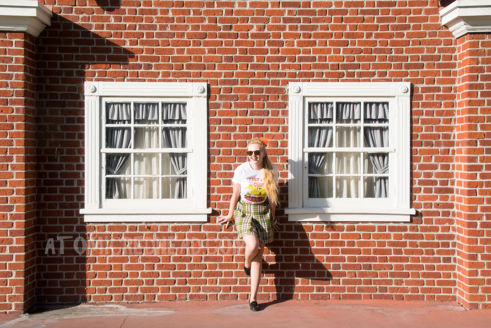 Myself, leaning against a brick wall, wearing a white t-shirt with an image of a green ghost in a witch hat, rising from a silhouette of a town. Orange text reads "I met the Ghost of Oakhaven (and lived)," a skirt in matching green, orange, white and black, and an orange scarf in my pony tail.