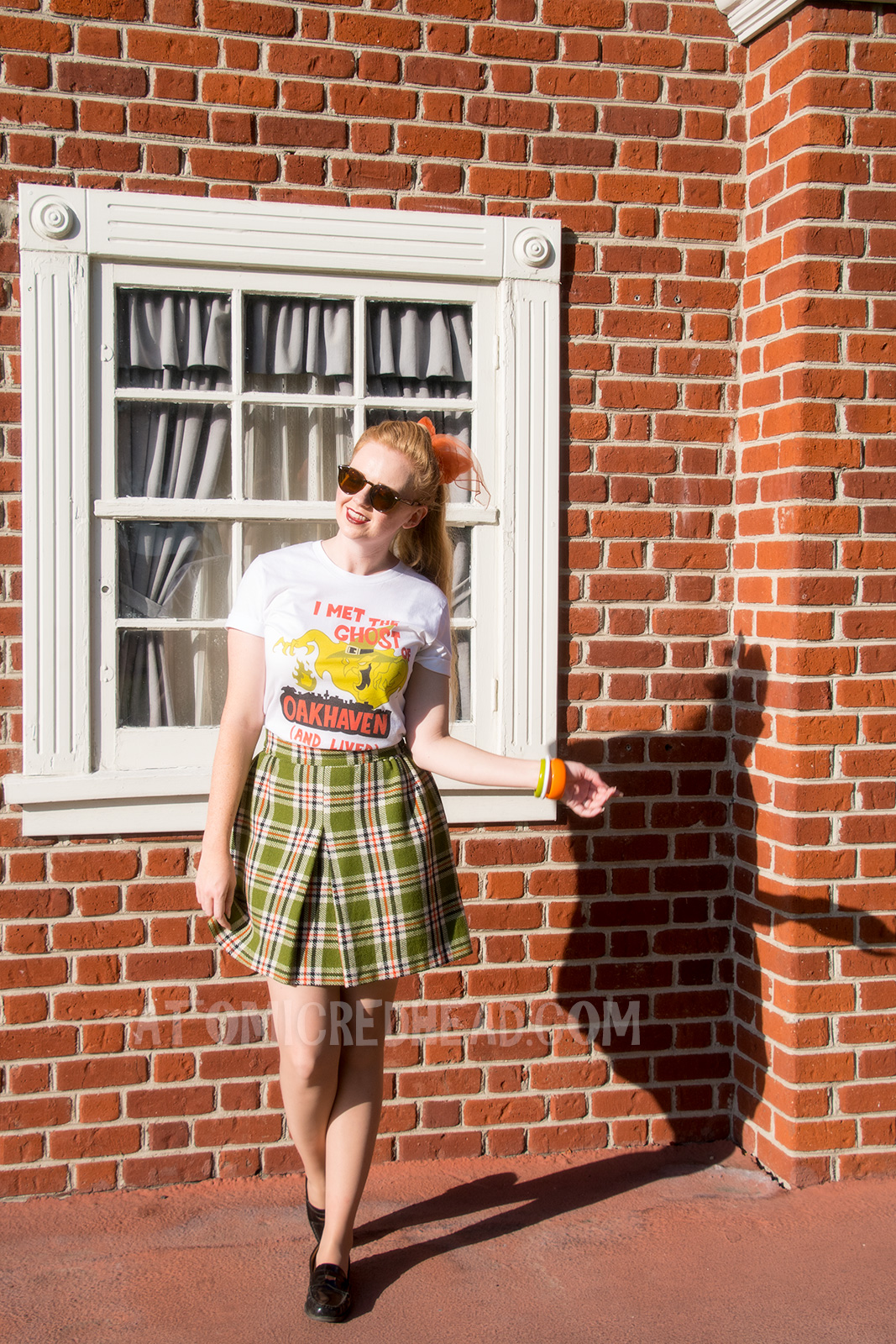 Myself, standing in front of a brick wall, wearing a white t-shirt with an image of a green ghost in a witch hat, rising from a silhouette of a town. Orange text reads "I met the Ghost of Oakhaven (and lived)," a skirt in matching green, orange, white and black, and an orange scarf in my pony tail.