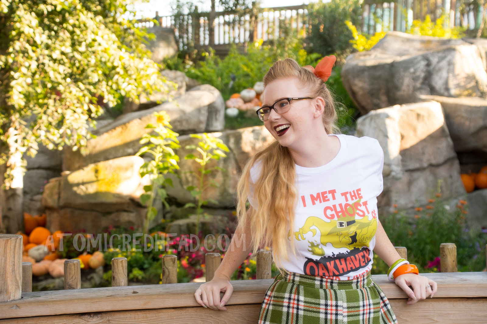 Myself, standing in front of a small lake with cascading waterfall in the background and pumpkins scattered about, wearing a white t-shirt with an image of a green ghost in a witch hat, rising from a silhouette of a town. Orange text reads "I met the Ghost of Oakhaven (and lived)," a skirt in matching green, orange, white and black, and an orange scarf in my pony tail.