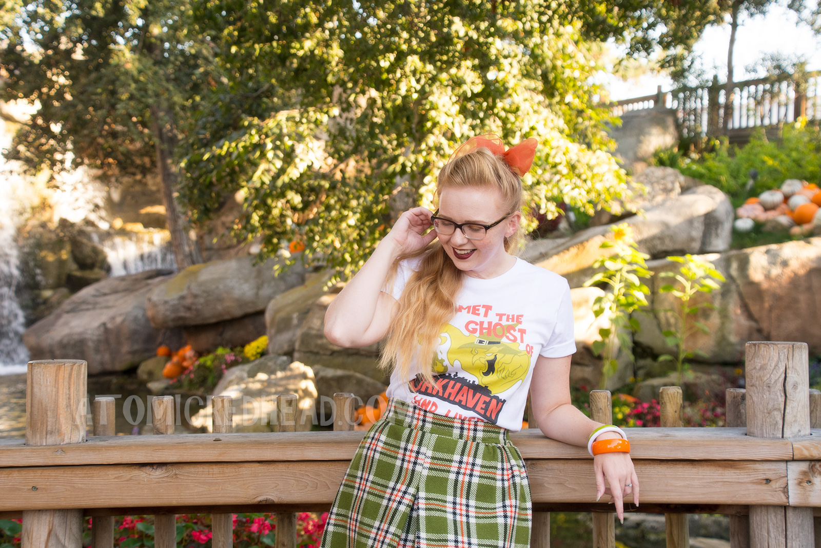 Myself, standing in front of a small lake with cascading waterfall in the background and pumpkins scattered about, wearing a white t-shirt with an image of a green ghost in a witch hat, rising from a silhouette of a town. Orange text reads "I met the Ghost of Oakhaven (and lived)," a skirt in matching green, orange, white and black, and an orange scarf in my pony tail.