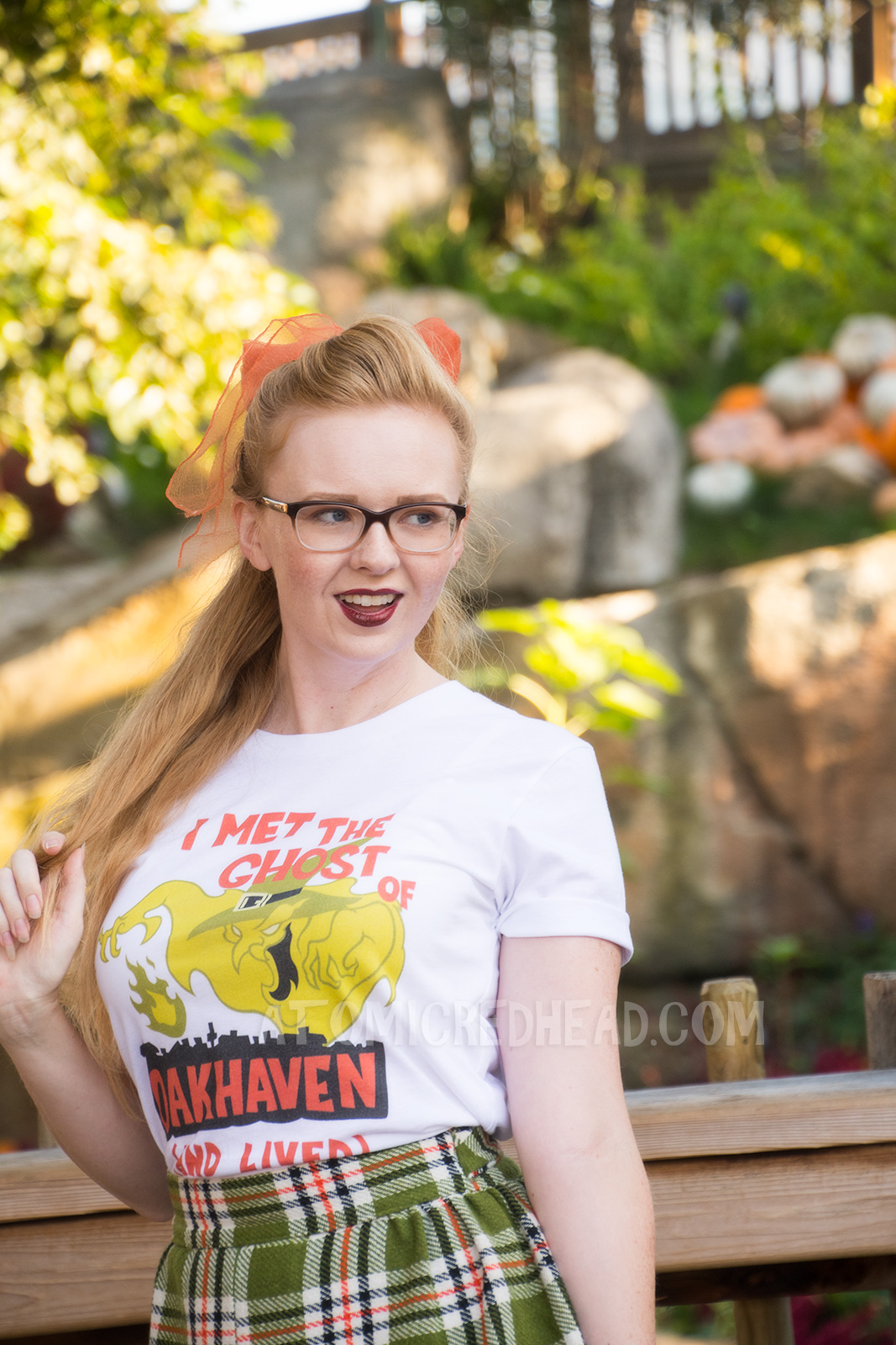 Myself, standing in front of a small lake with cascading waterfall in the background and pumpkins scattered about, wearing a white t-shirt with an image of a green ghost in a witch hat, rising from a silhouette of a town. Orange text reads "I met the Ghost of Oakhaven (and lived)," a skirt in matching green, orange, white and black, and an orange scarf in my pony tail.