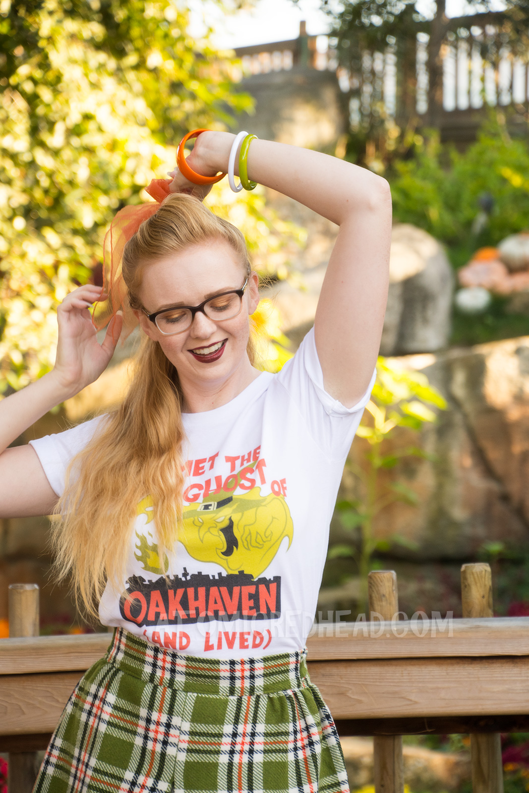 Myself, standing in front of a small lake with cascading waterfall in the background and pumpkins scattered about, wearing a white t-shirt with an image of a green ghost in a witch hat, rising from a silhouette of a town. Orange text reads "I met the Ghost of Oakhaven (and lived)," a skirt in matching green, orange, white and black, and an orange scarf in my pony tail.