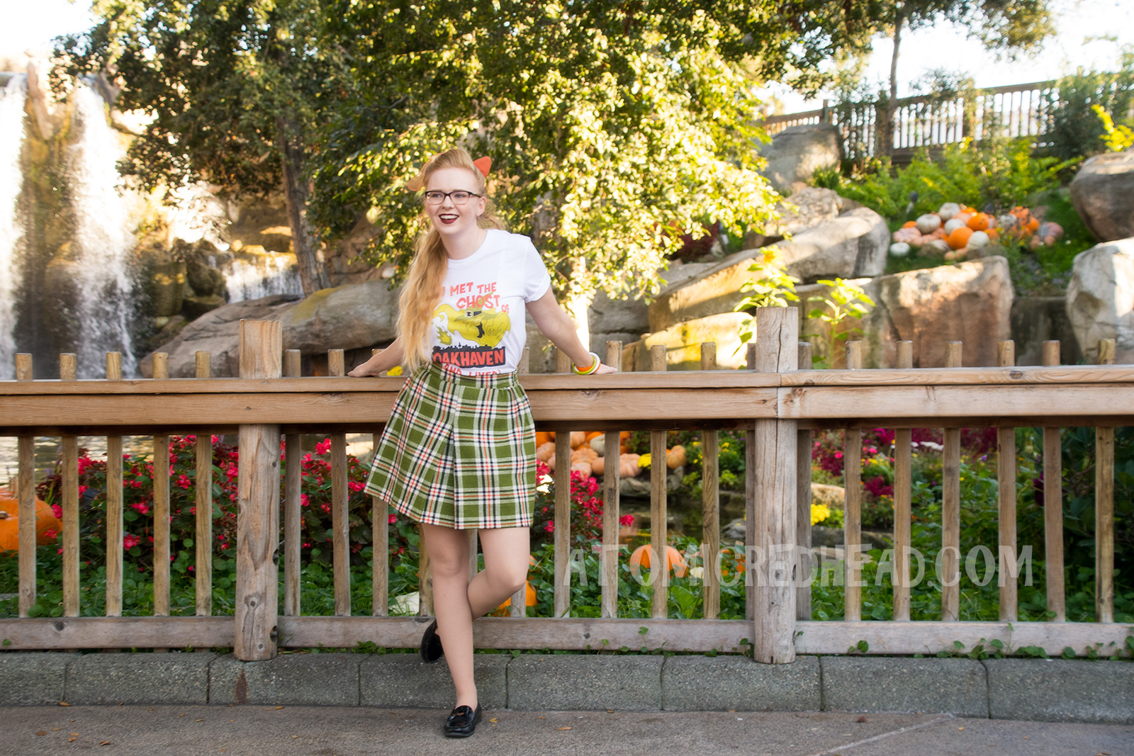 Myself, standing in front of a small lake with cascading waterfall in the background and pumpkins scattered about, wearing a white t-shirt with an image of a green ghost in a witch hat, rising from a silhouette of a town. Orange text reads "I met the Ghost of Oakhaven (and lived)," a skirt in matching green, orange, white and black, and an orange scarf in my pony tail.