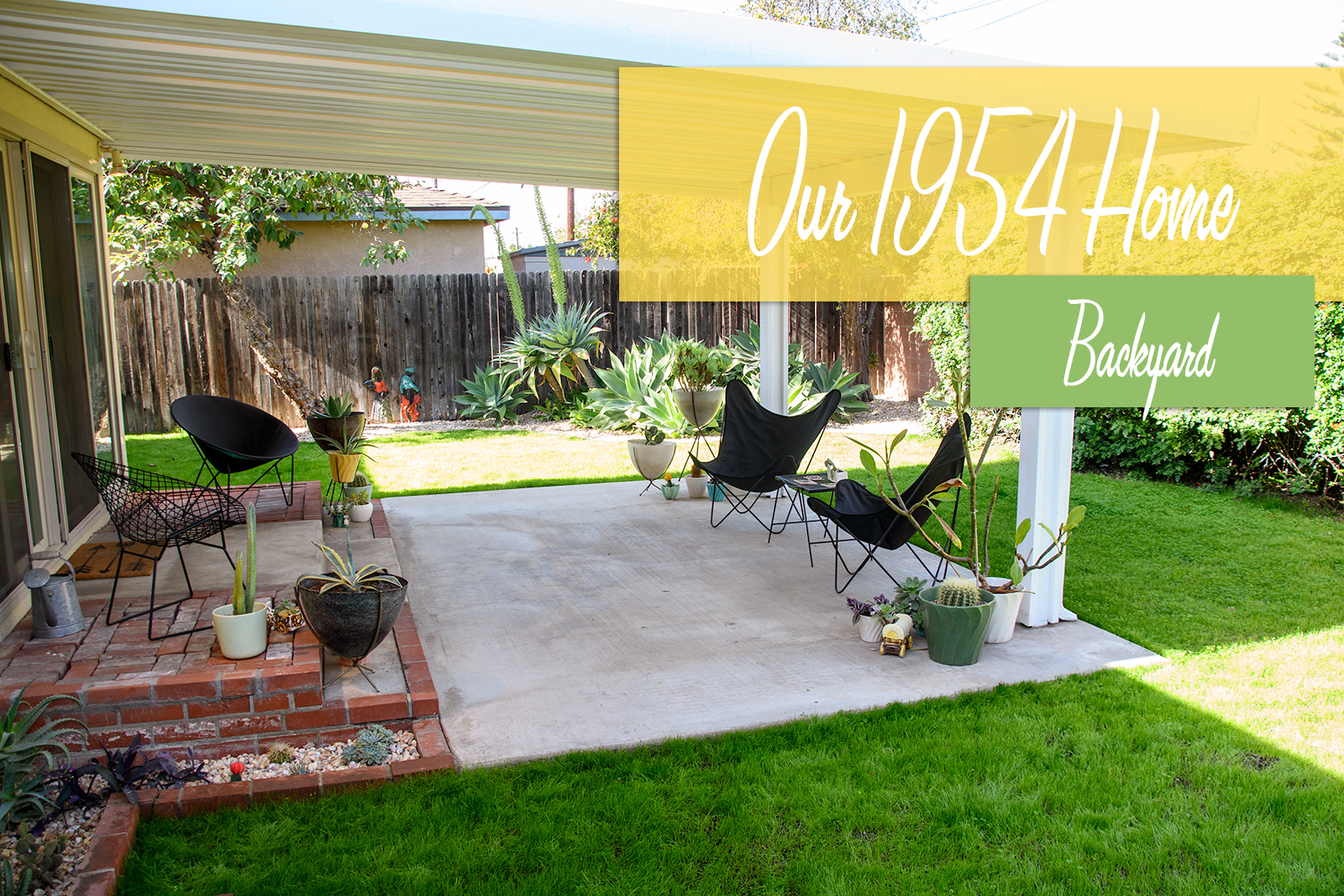 A covered patio stretches out from a butter yellow house, on the patio are various ceramic and fiberglass planters, along with two black butterfly style chairs. Green grass surrounds the patio, large agave plants are clustered in the corner.