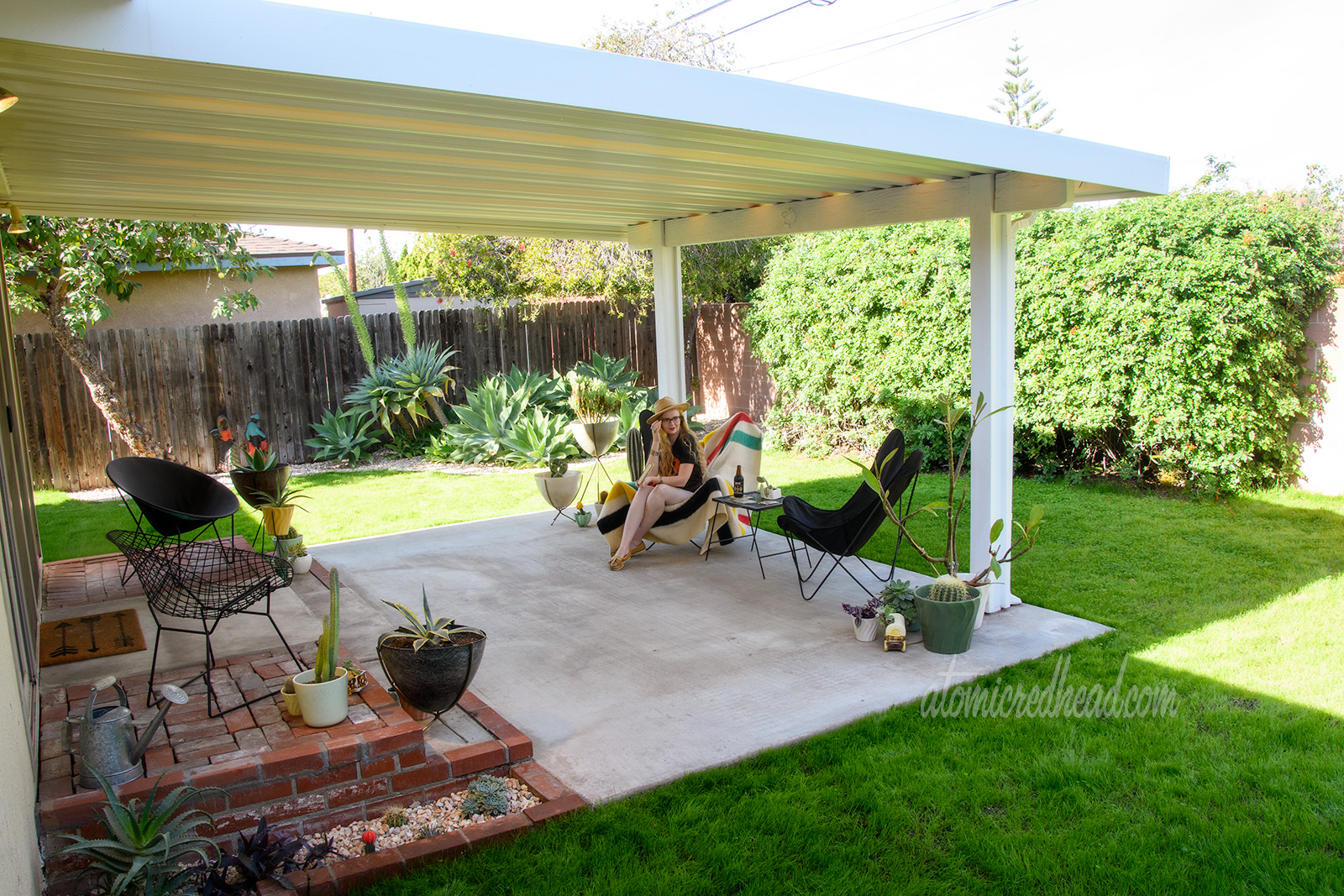 A covered patio stretches out from a butter yellow house, on the patio are various ceramic and fiberglass planters, along with two black butterfly style chairs. Green grass surrounds the patio, large agave plants are clustered in the corner. I am seated in one of the butterfly chairs reading a book, wearing a black t-shirt featuring a jackalope, and a straw hat.