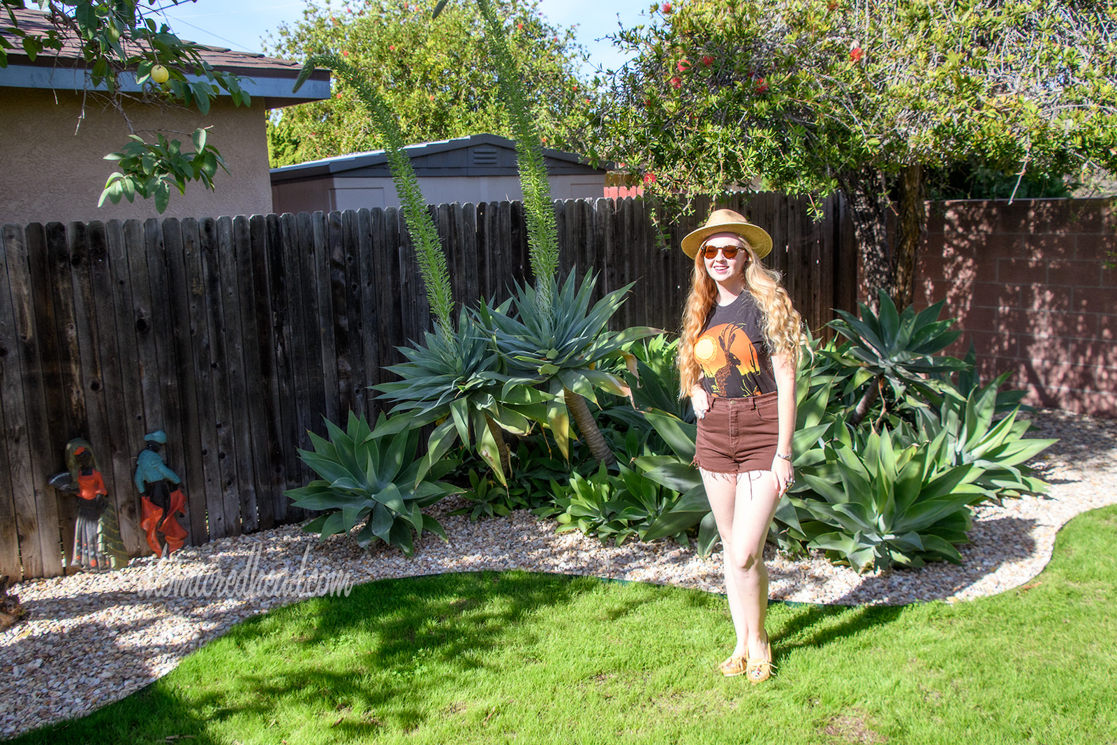 Myself standing in front of the agave plants, wearing a black t-shirt with a jackalope on it, a pair of brown shorts, and a woven straw hat.