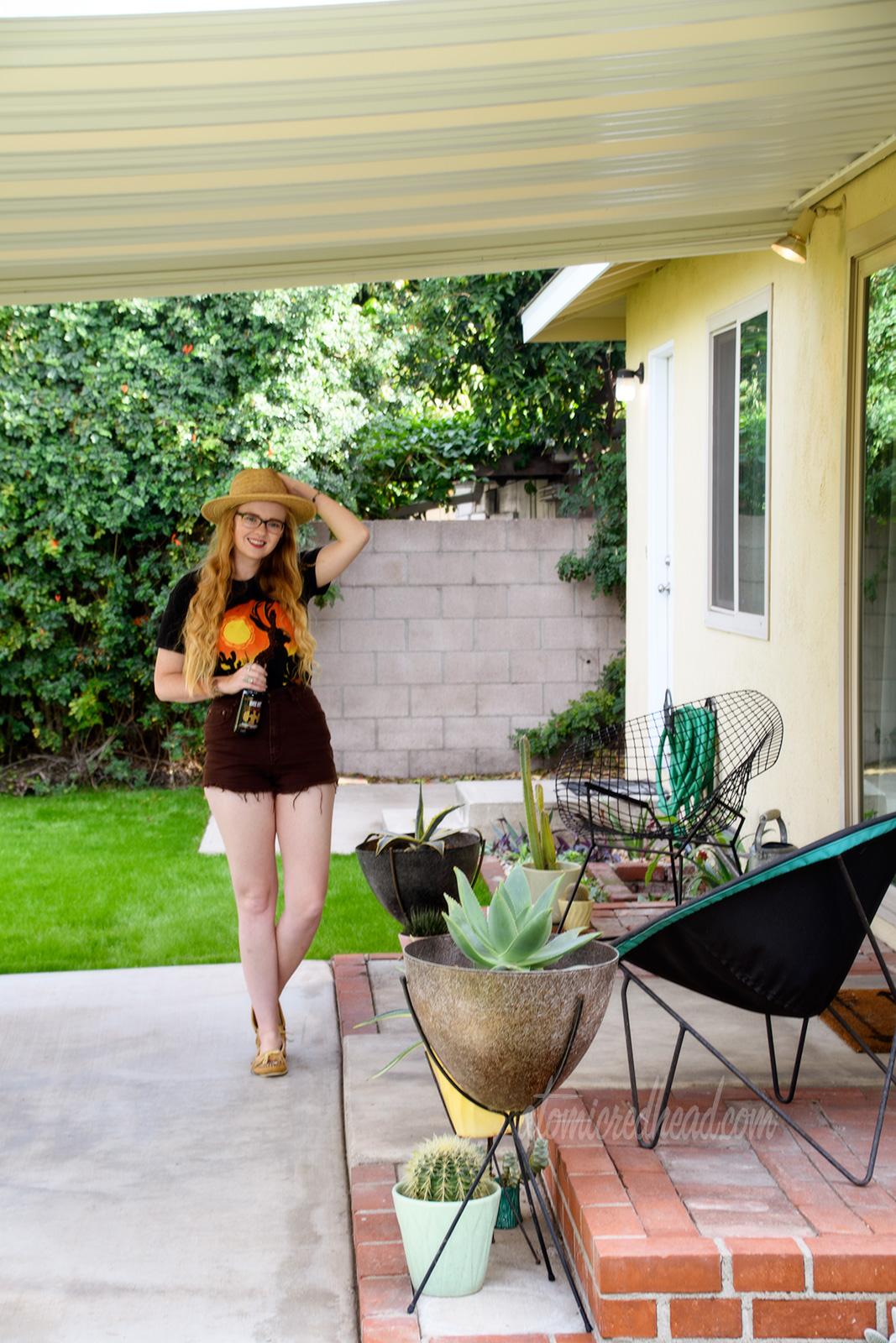 Myself standing on our patio, wearing a black t-shirt with a jackalope on it, brown shirts, and a woven straw hat. Planters scatter the patio with various cacti and succulants.