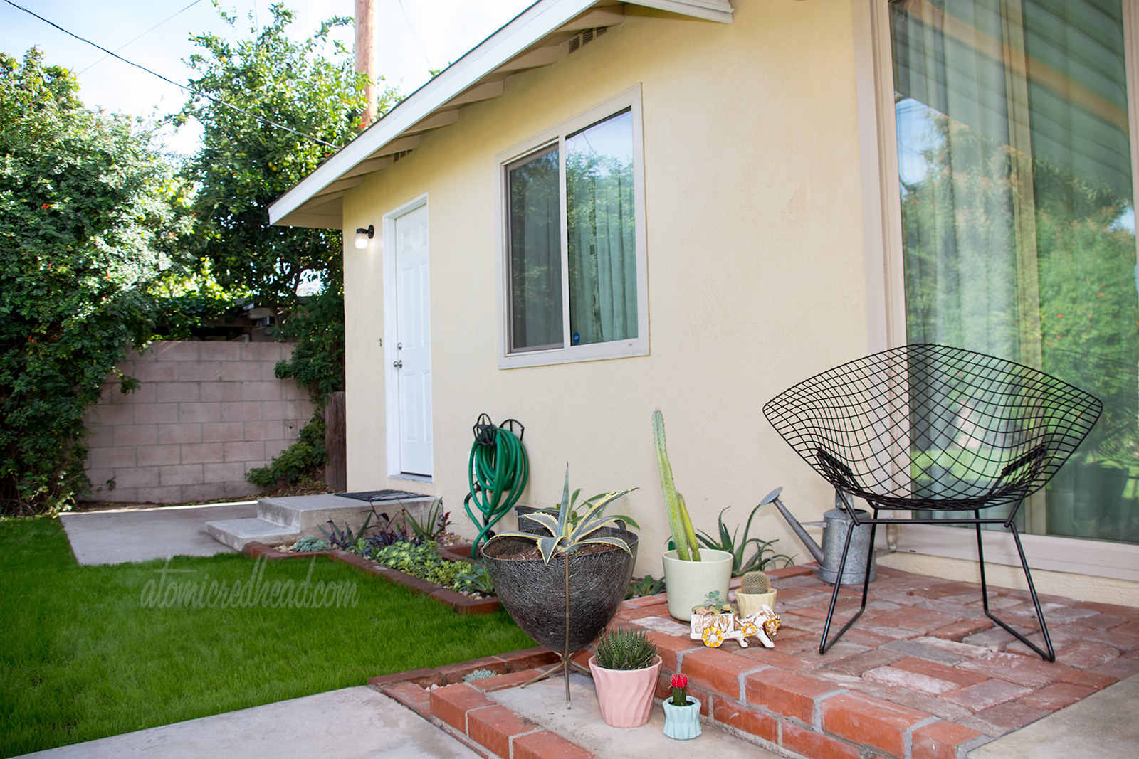 A wire diamond shaped chair sits on the patio, surrounded by various planters with cacti and succulents in them.