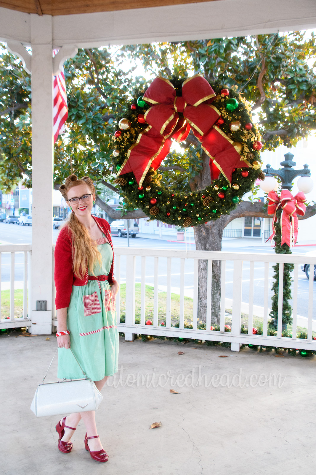 Myself standing in a covered white bandstand wearing a mint green dress with red and white stripe pockets, a red sweater, a Christmas tree brooch, white purse, and red shoes. A large Christmas wreath hangs in the background.