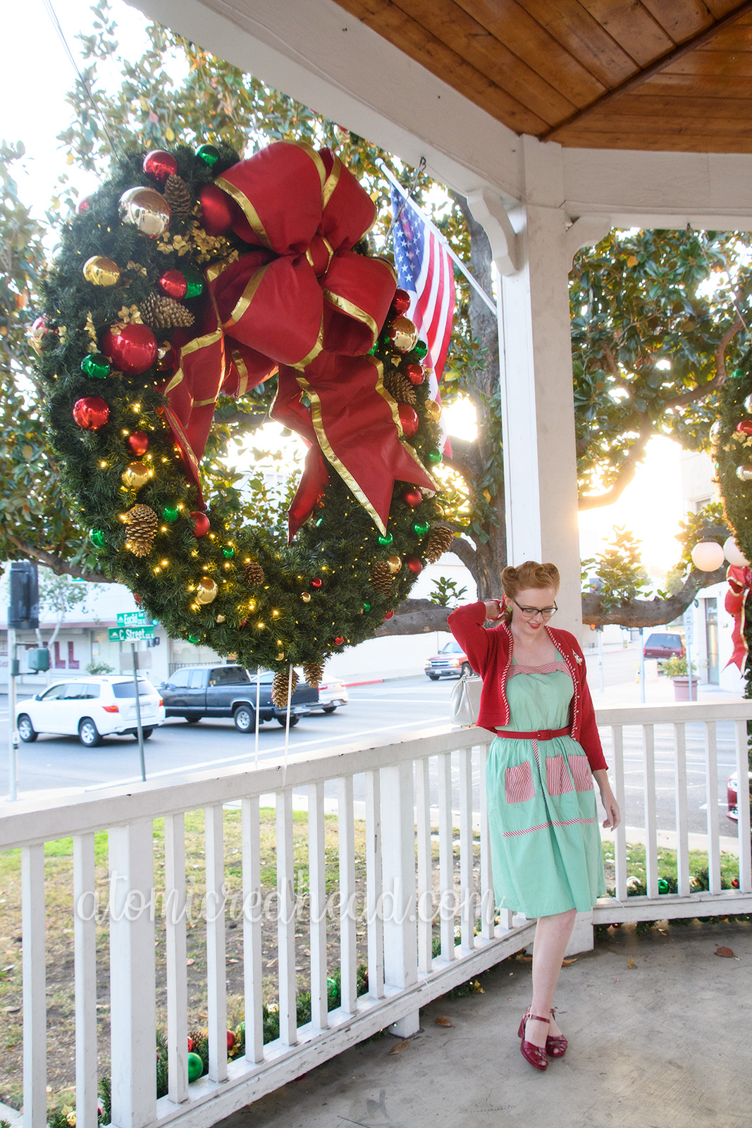 Myself standing in a covered white bandstand wearing a mint green dress with red and white stripe pockets, a red sweater, a Christmas tree brooch, white purse, and red shoes. A large Christmas wreath hangs in the background.