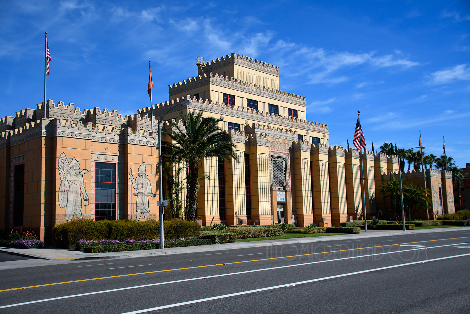 A three story imposing looking building stands, it is blocky, and tiers upwards, and in shades of yellow, pale orange, and tan.