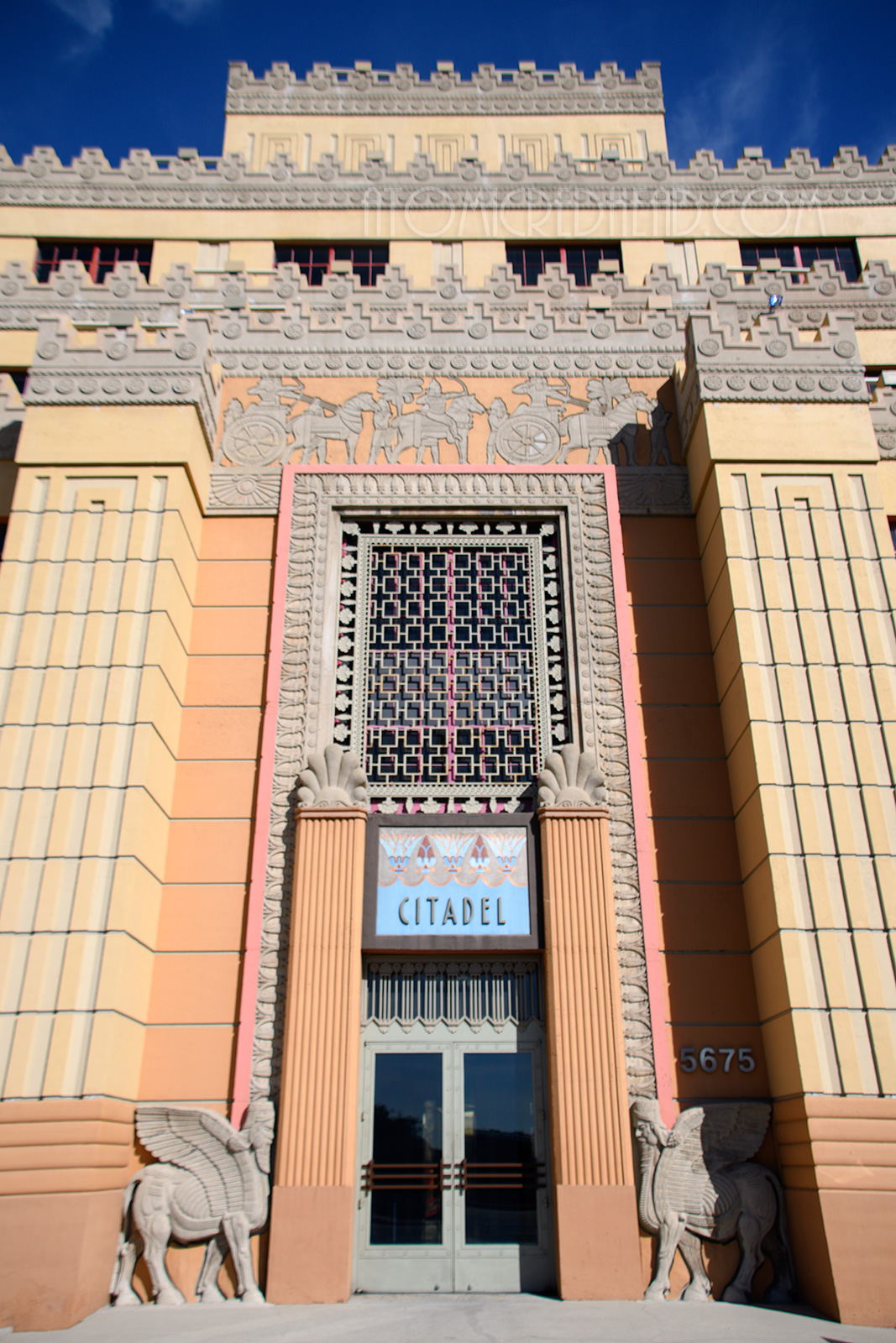 The front doors to the Citadel sit between tall columns, on either side Lamassu, winged men with four cloven feet, flank the doors. Above the door reads "Citadel" with lotus flowers above. Along the top are carvings of chariot races.