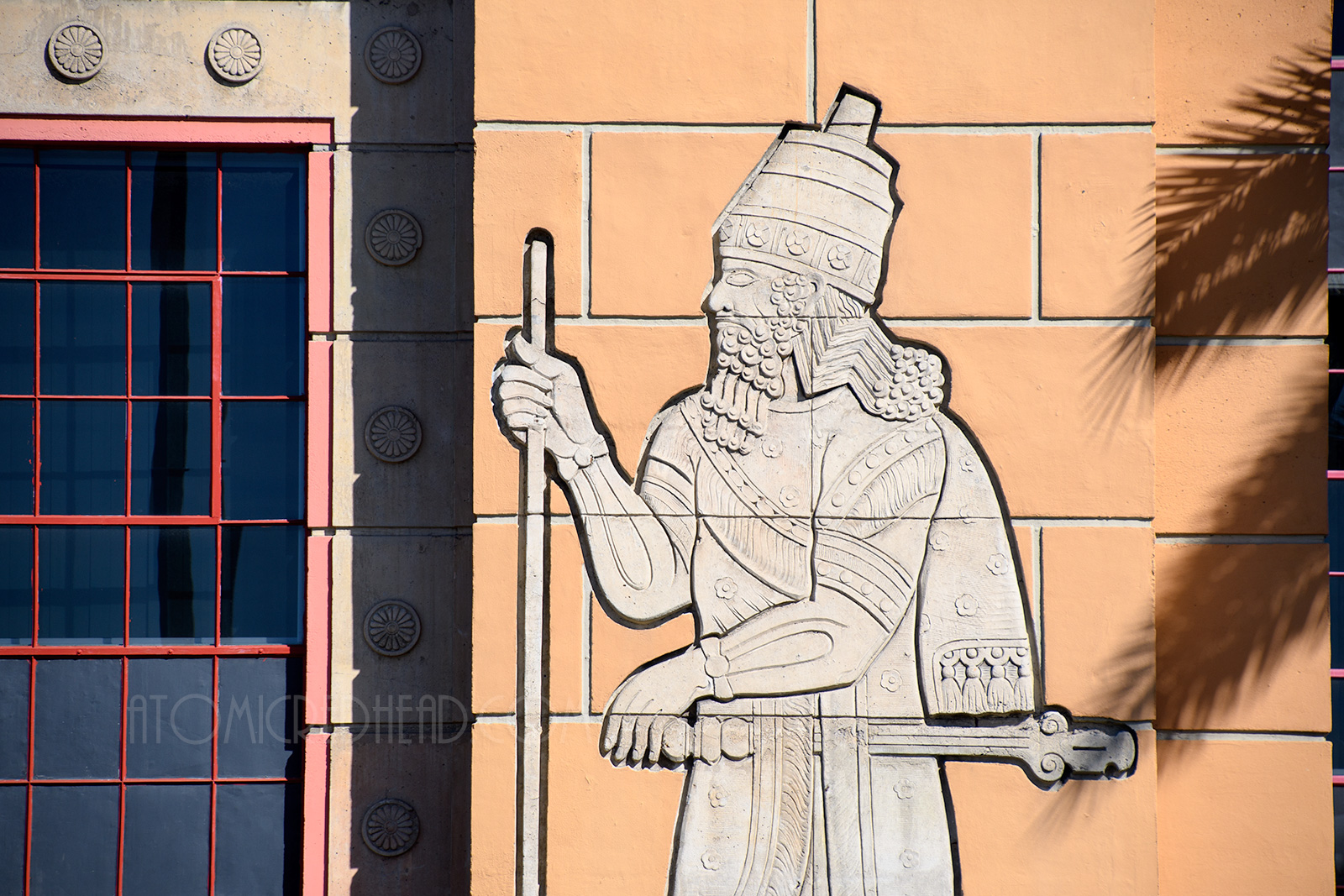 Close-up of the carving of King Sargon, holding a staff, he has a beard, a tall crown like hat, and robes with elegant designs.