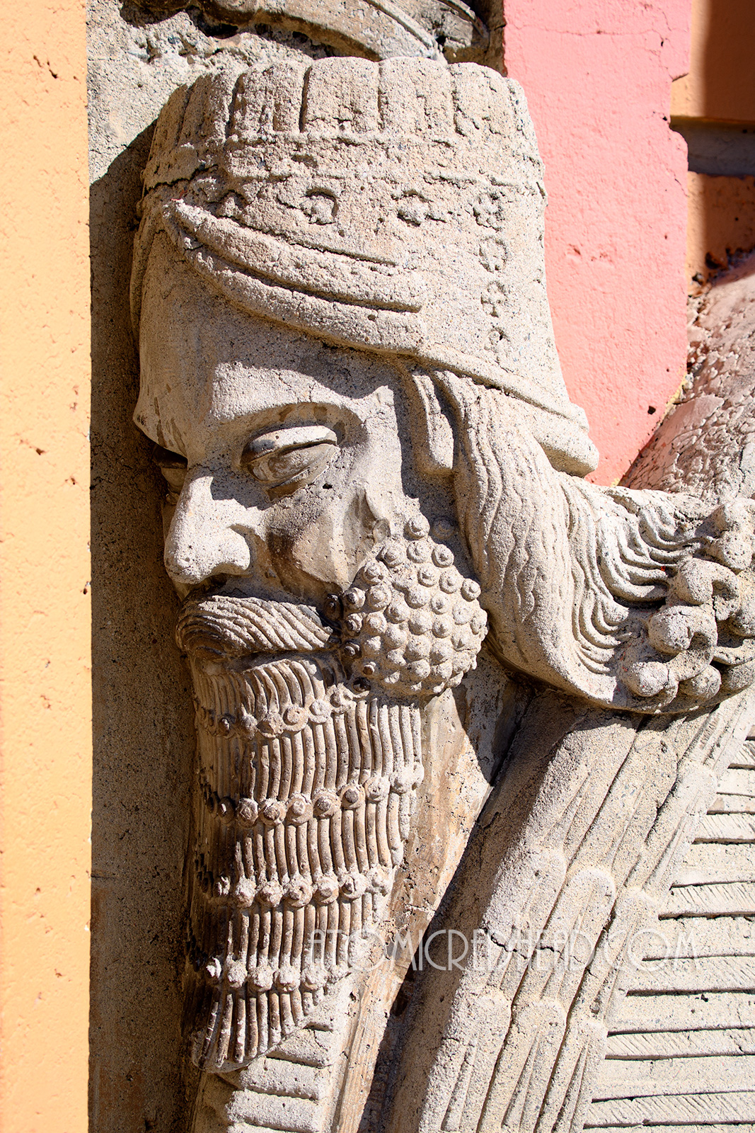 Close-up of the face of one of the Lamassu creatures that sit outside the door. He has a tall crown like hat and beard, and curly long hair.